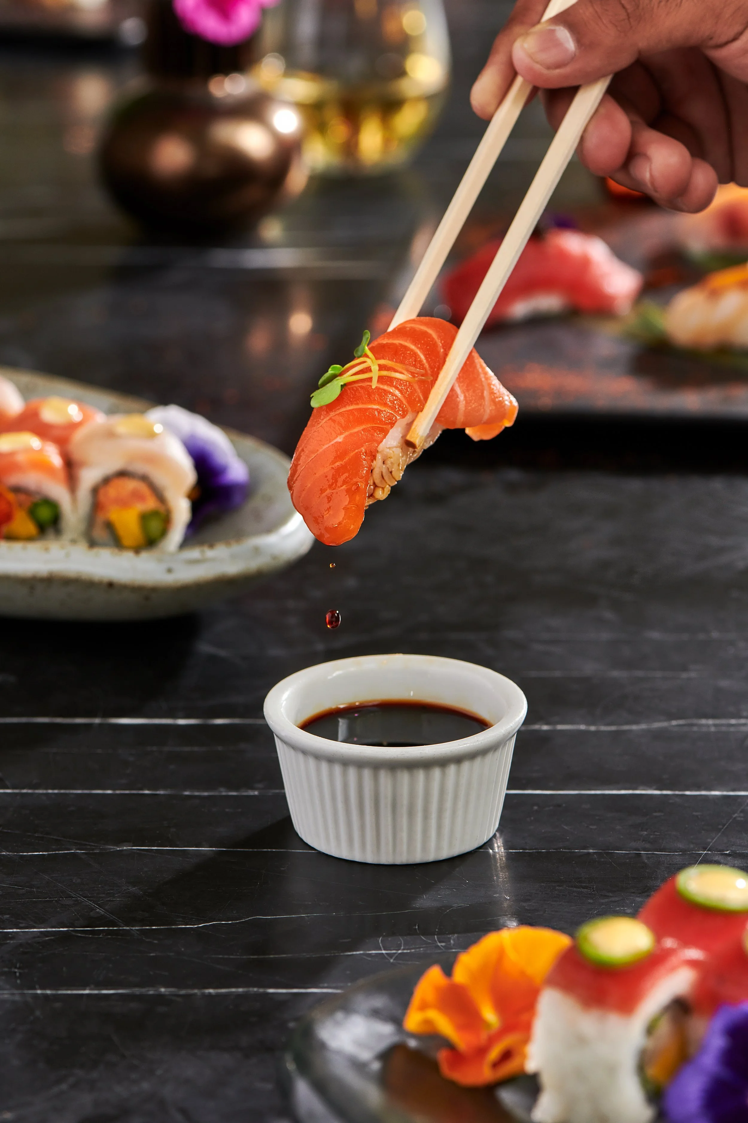 Person holding chopsticks with a piece of salmon sushi over a small white dish of soy sauce on a dark tabletop, with other sushi rolls in the background.