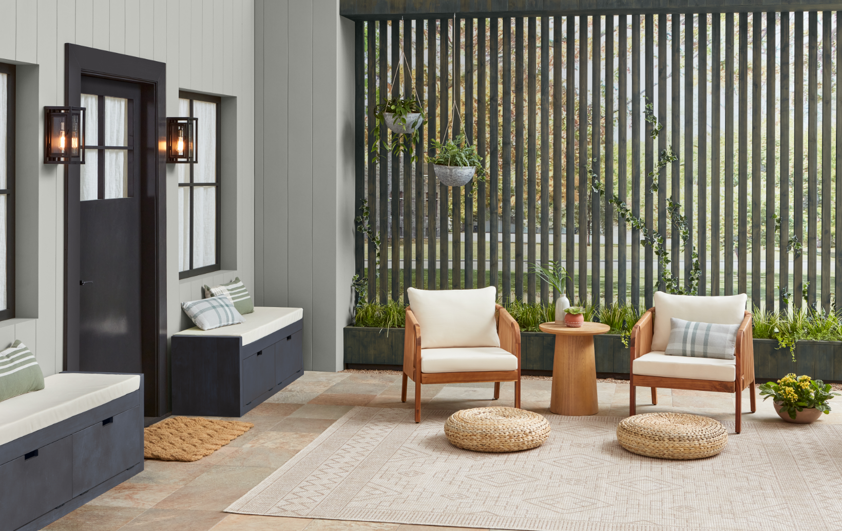 Interior of a modern patio with two white cushioned chairs, a small round wooden table, hanging planters, potted plants, and a decoratively designed rug. The background features a vertical wooden slat fence with greenery.
