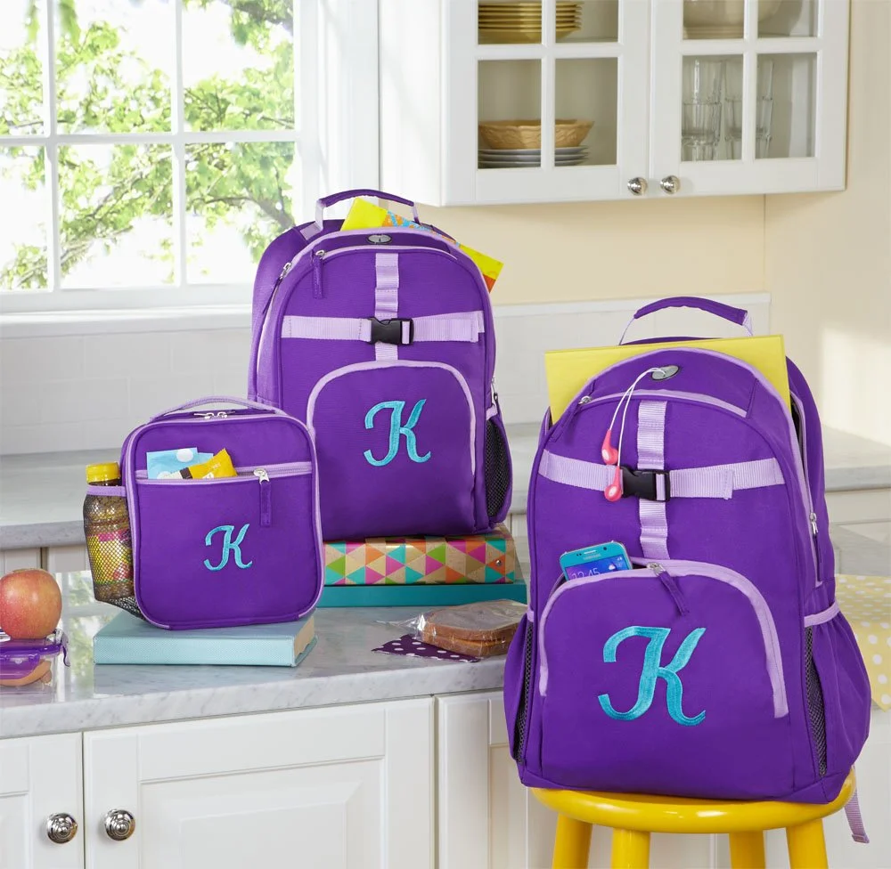 Four purple backpacks with light blue letter K on a white kitchen counter and yellow barstool. One backpack has pink earbuds, another has a smartphone. The backpacks contain yellow folders and snacks. A small purple lunchbox with a yellow water bottl