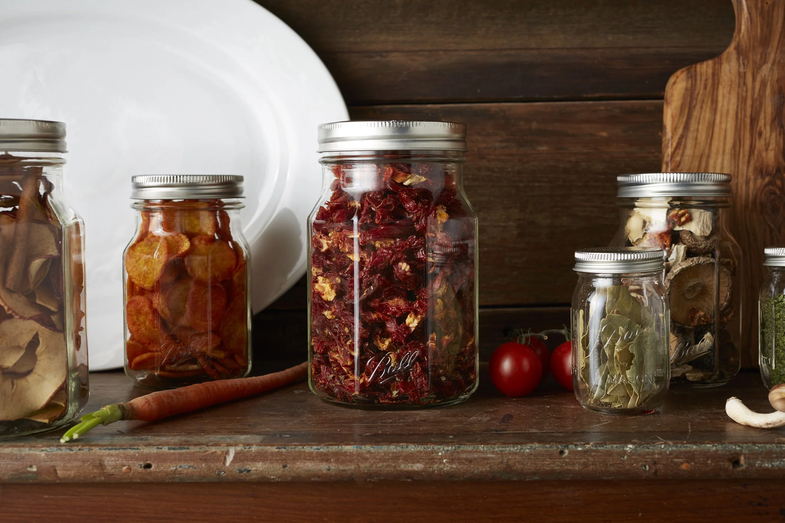 Assorted dried herbs, vegetables, and spices in glass jars on a wooden shelf, with fresh cherry tomatoes and a carrot in front.
