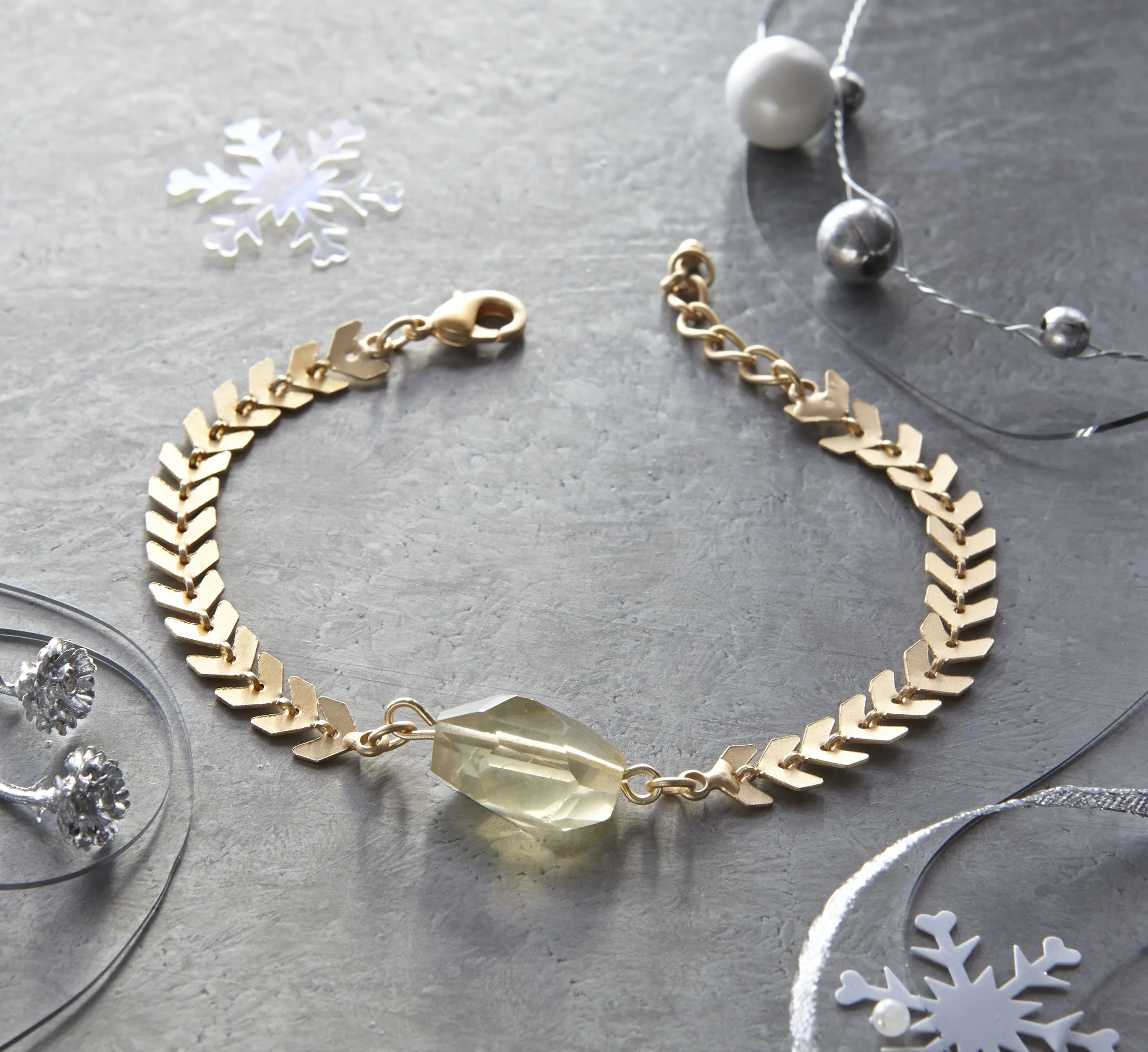 Gold bracelet with a large yellowish gemstone resting on a gray surface, surrounded by silver and white holiday decorations including a snowflake, pearls, and metallic spheres.