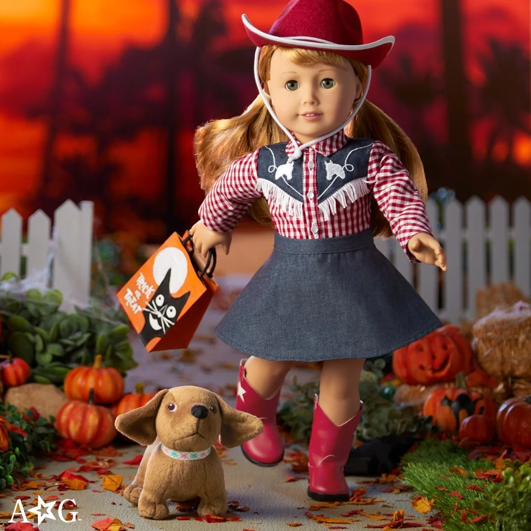 An American Girl doll dressed in a cowboy-themed outfit, wearing a red cowboy hat, plaid shirt, denim skirt, and red cowboy boots, is standing outdoors on a fall-themed display with pumpkins, autumn leaves, and a small plush dog, during sunset.