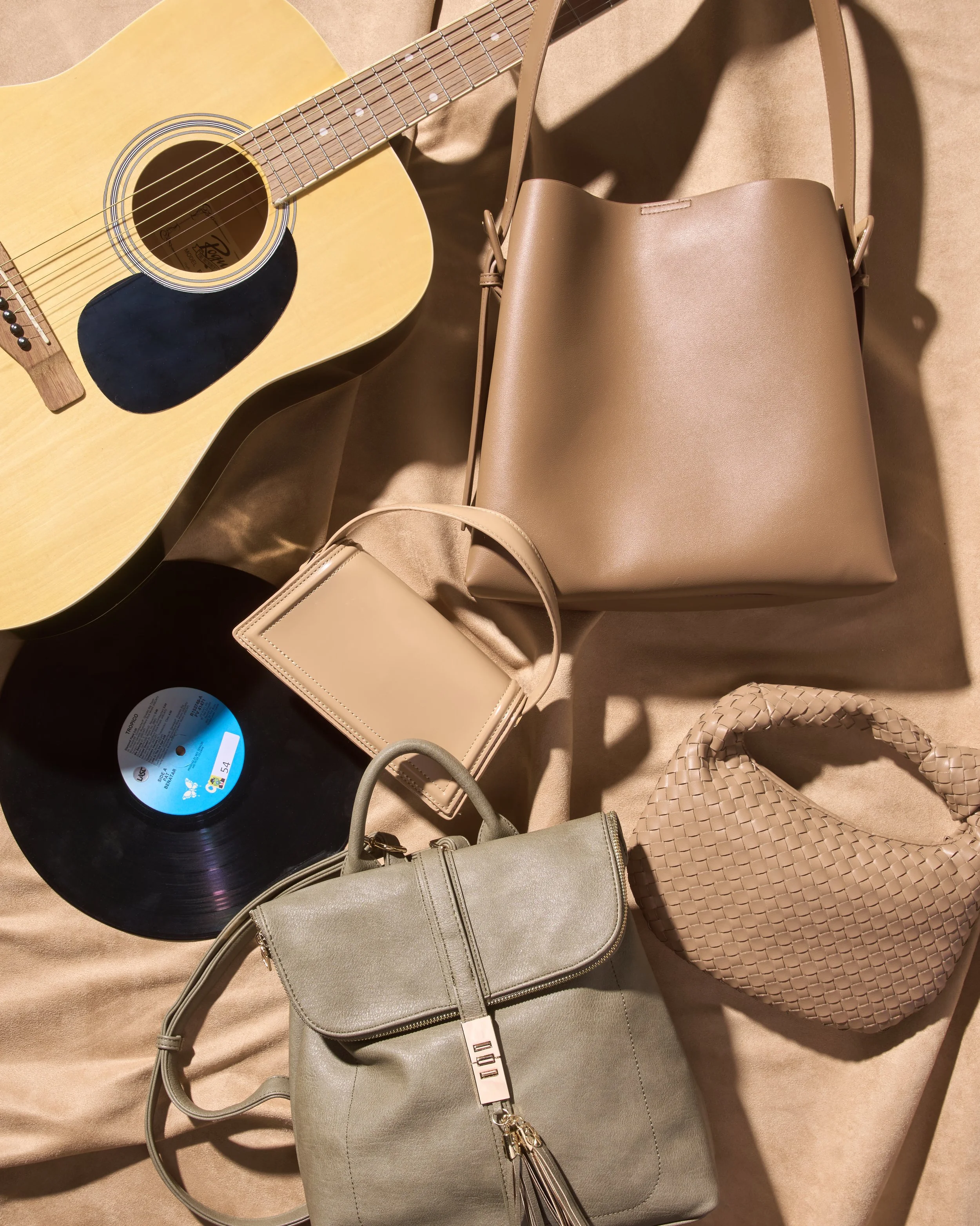 An acoustic guitar, a vinyl record, and multiple beige and gray handbags on a beige fabric surface.