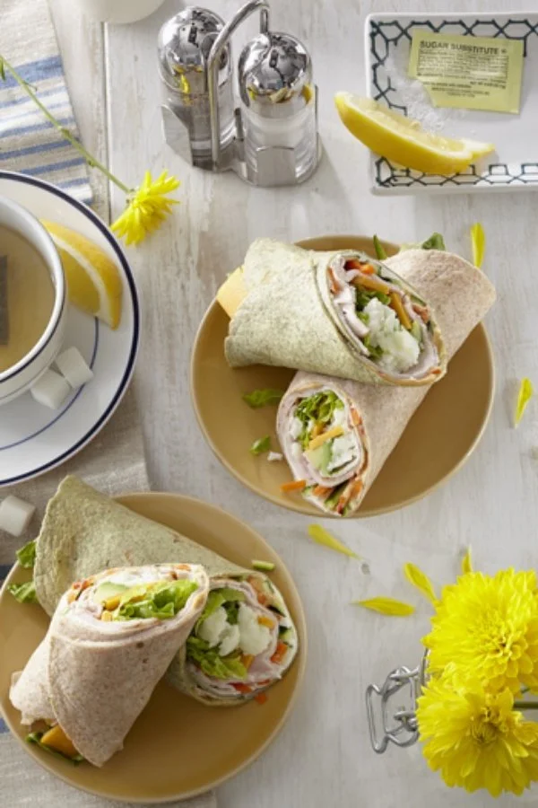 Two plates with turkey wrap sandwiches filled with lettuce, tomato, and cheese, beside a cup of tea with lemon and a salt and pepper shaker. A lemon wedge and yellow flowers on a white table.