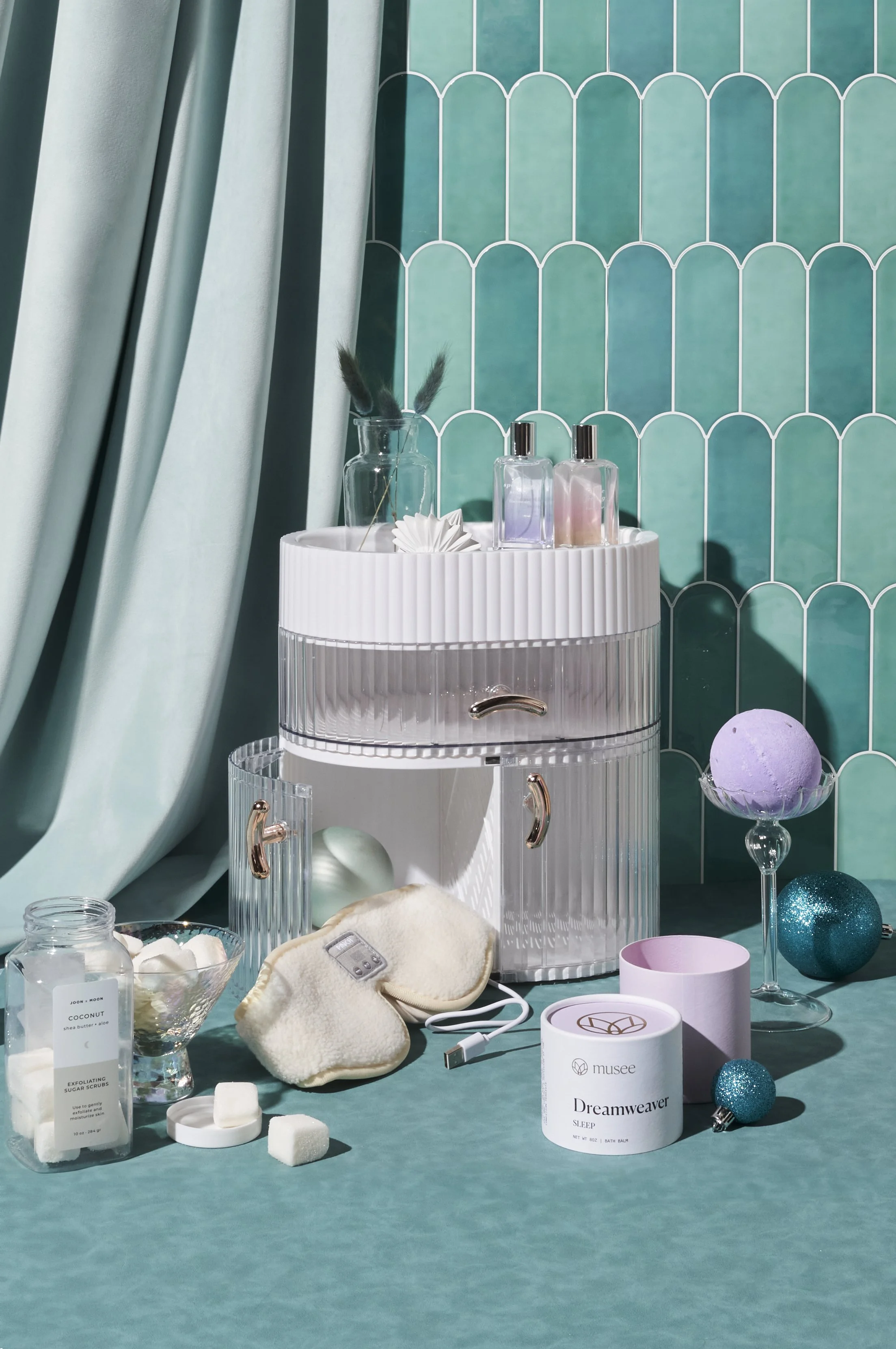 A collection of skincare and beauty products on a teal surface with teal curtains and wall tiles, including bath salts, glittery ornaments, candles, and a small storage unit.