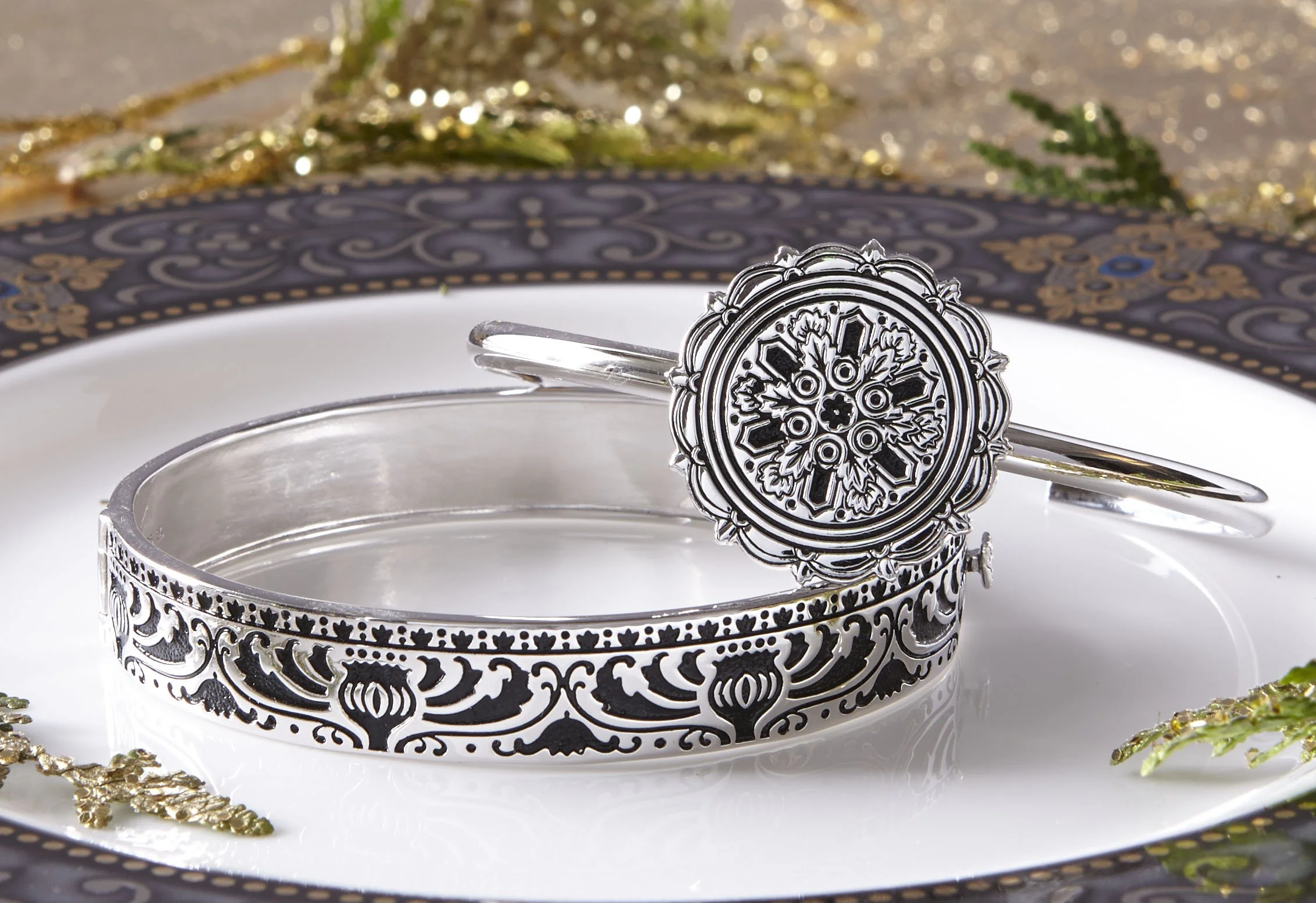 Silver jewelry including a bangle and a bracelet with intricate black and silver designs, displayed on a black and gold plate with decorative edges, with gold and green embellishments in the background.