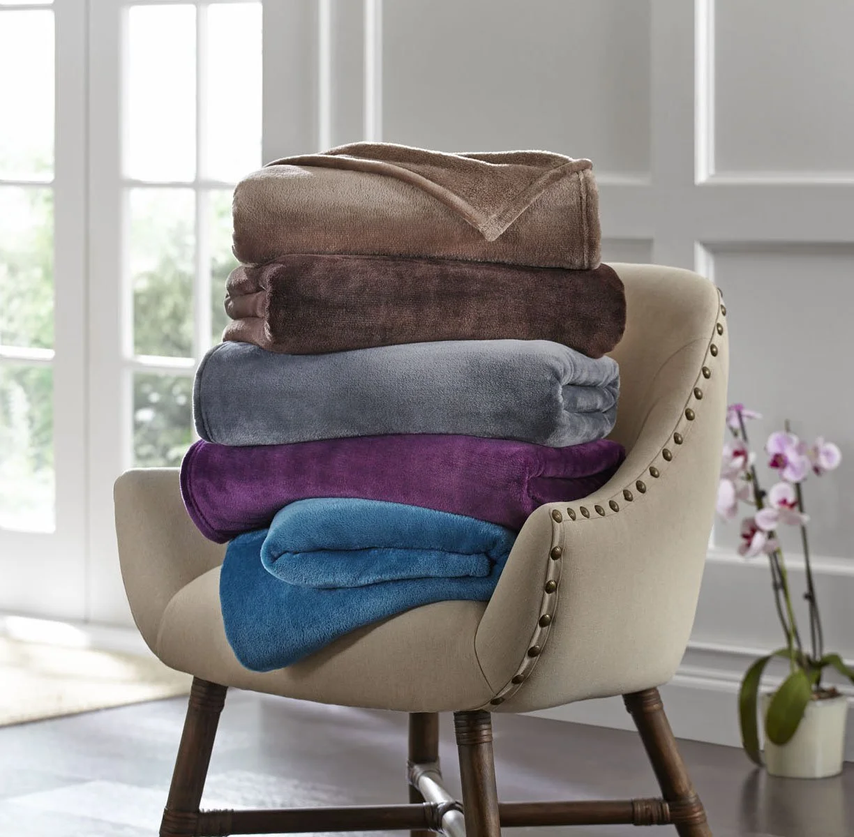 A beige upholstered chair with dark wooden legs, filled with a tall stack of colorful folded blankets or throws, in front of a window with white paneled doors and pink orchids on the side.