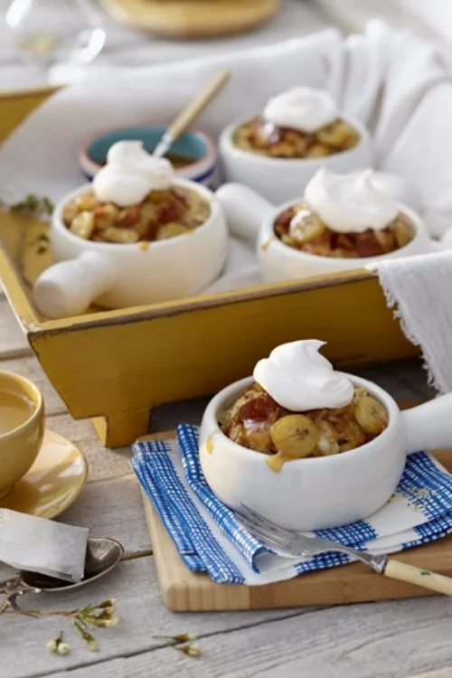 Four individual baked fruit cobblers topped with whipped cream, served in small white ramekins on a rustic wooden table with a blue and white cloth napkin and a silver spoon, with a yellow wooden tray in the background holding additional ramekins.
