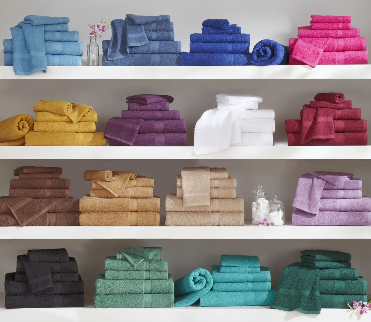 Colorful towels neatly folded and stacked on white shelves, organized by color, with small decorative flowers and jars in between.
