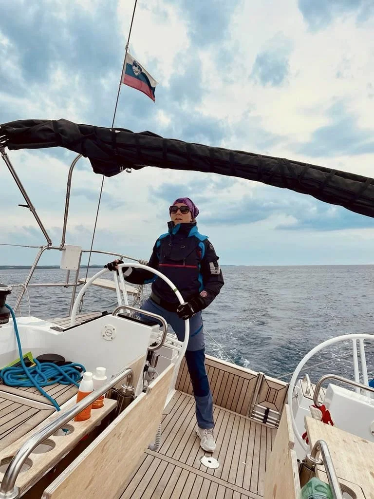 Alexandra Pichler steht auf einem Segelschiff auf dem Meer, trägt eine Jacke, Sonnenbrille und ein Kopftuch, hält das Steuerrad, im Hintergrund Himmel und Wasser.
