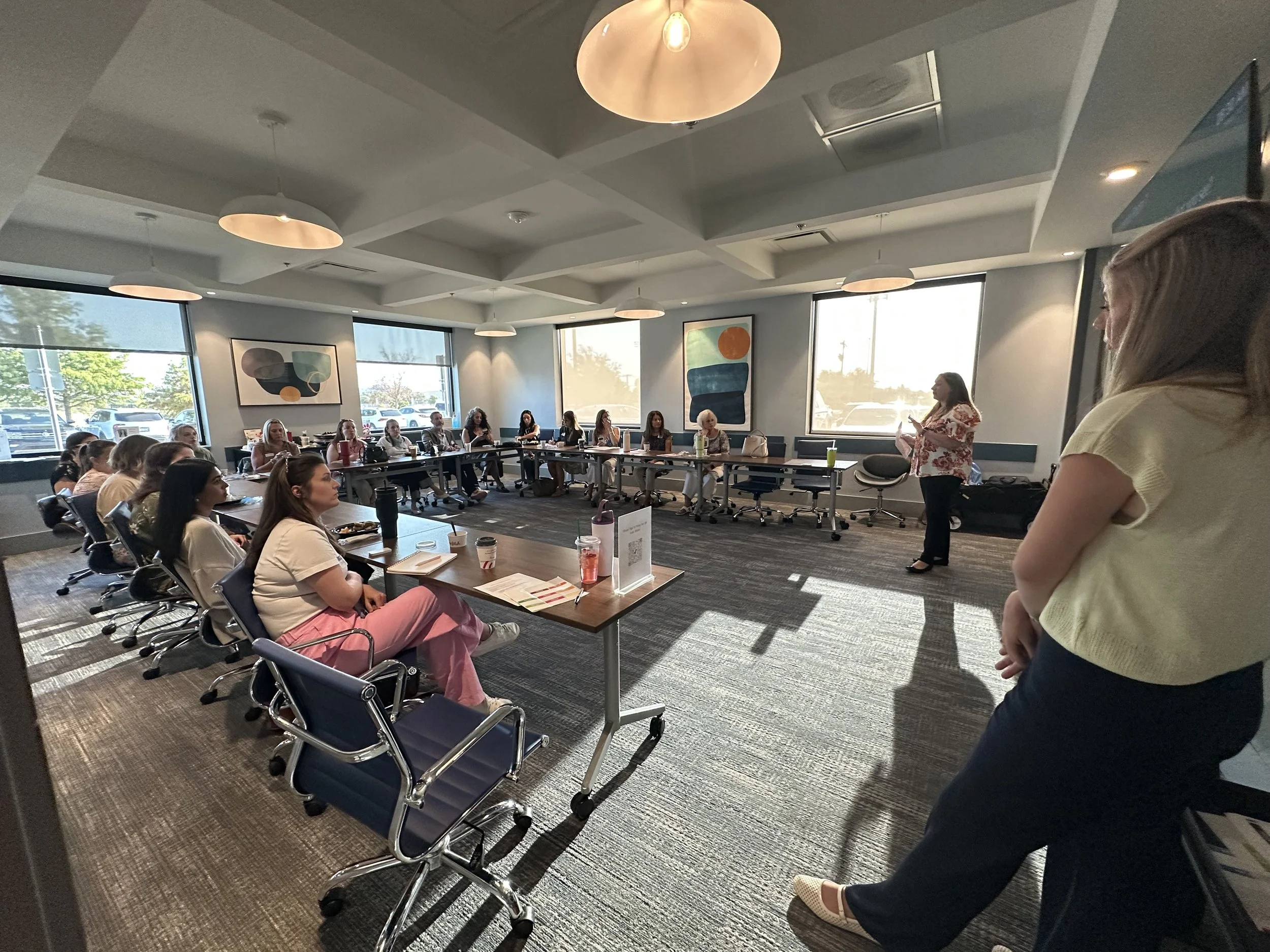 A group of people attending a meeting or presentation in a conference room with large windows, abstract artwork on the walls, and a woman standing and speaking.
