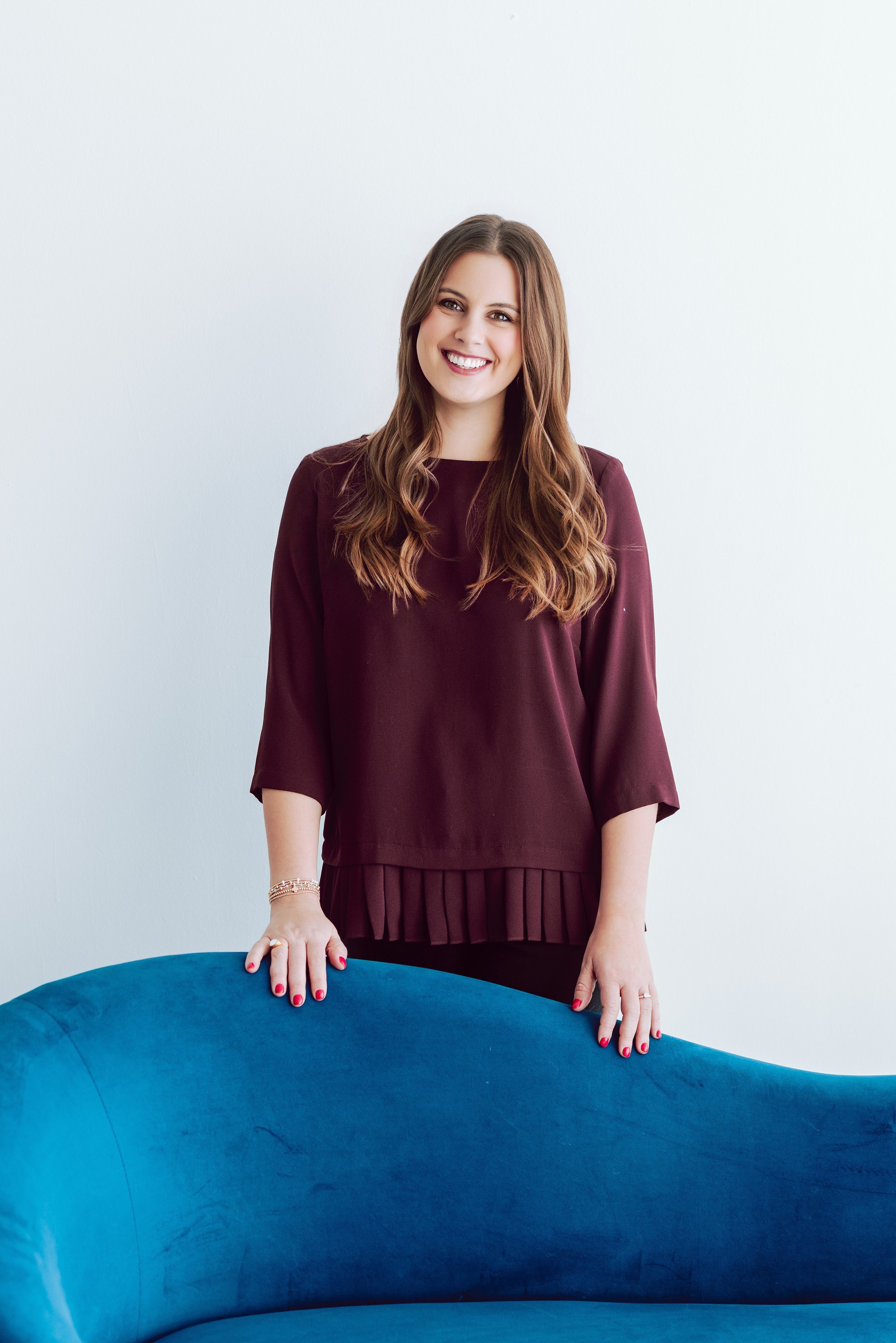 A woman with long, wavy brown hair and a bright smile, standing behind a blue velvet couch against a plain white wall, wearing a maroon top with pleated hem and red nail polish.