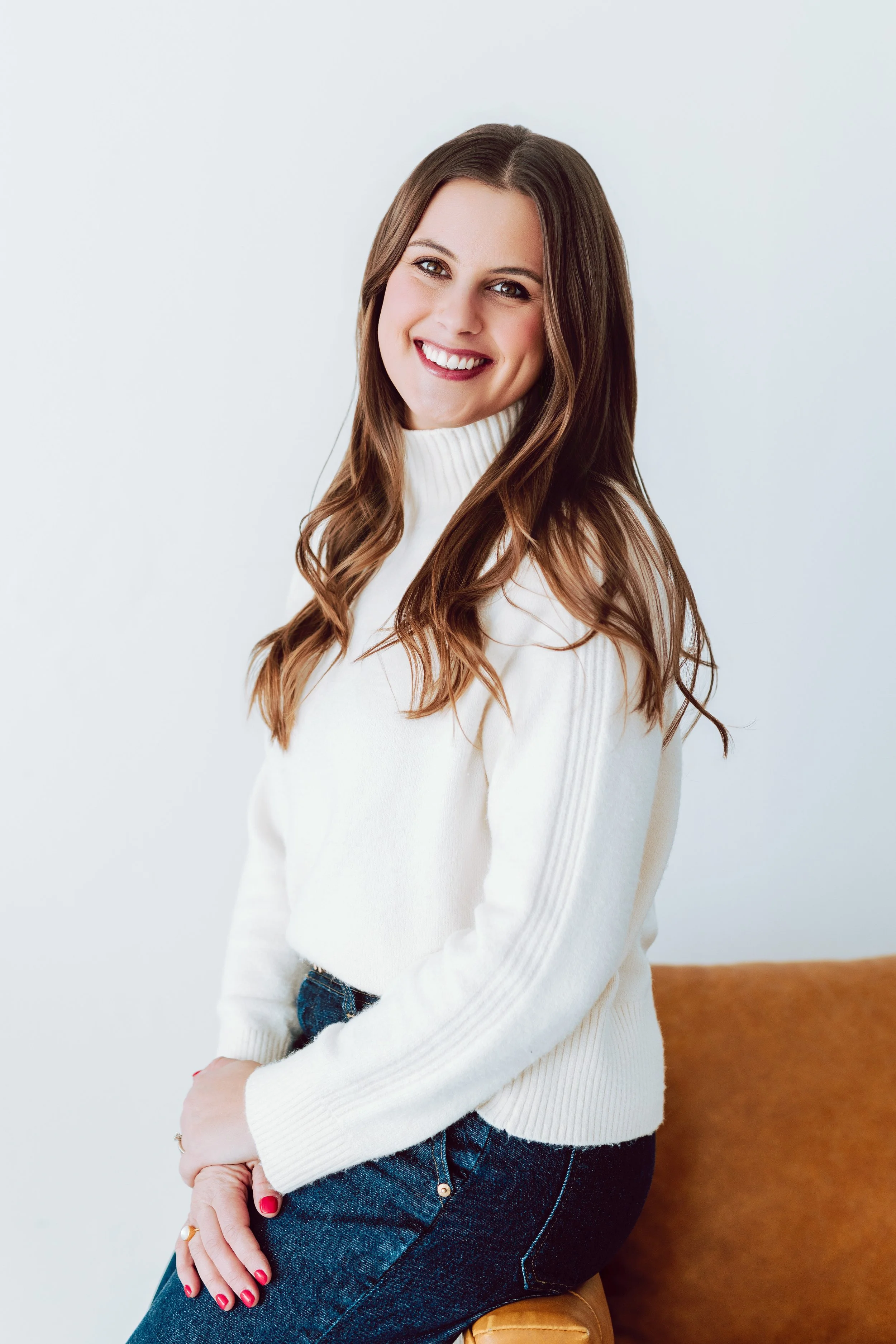 A smiling woman with long brown hair wearing a white turtleneck sweater and dark jeans, sitting on a brown chair or stool against a plain white background.