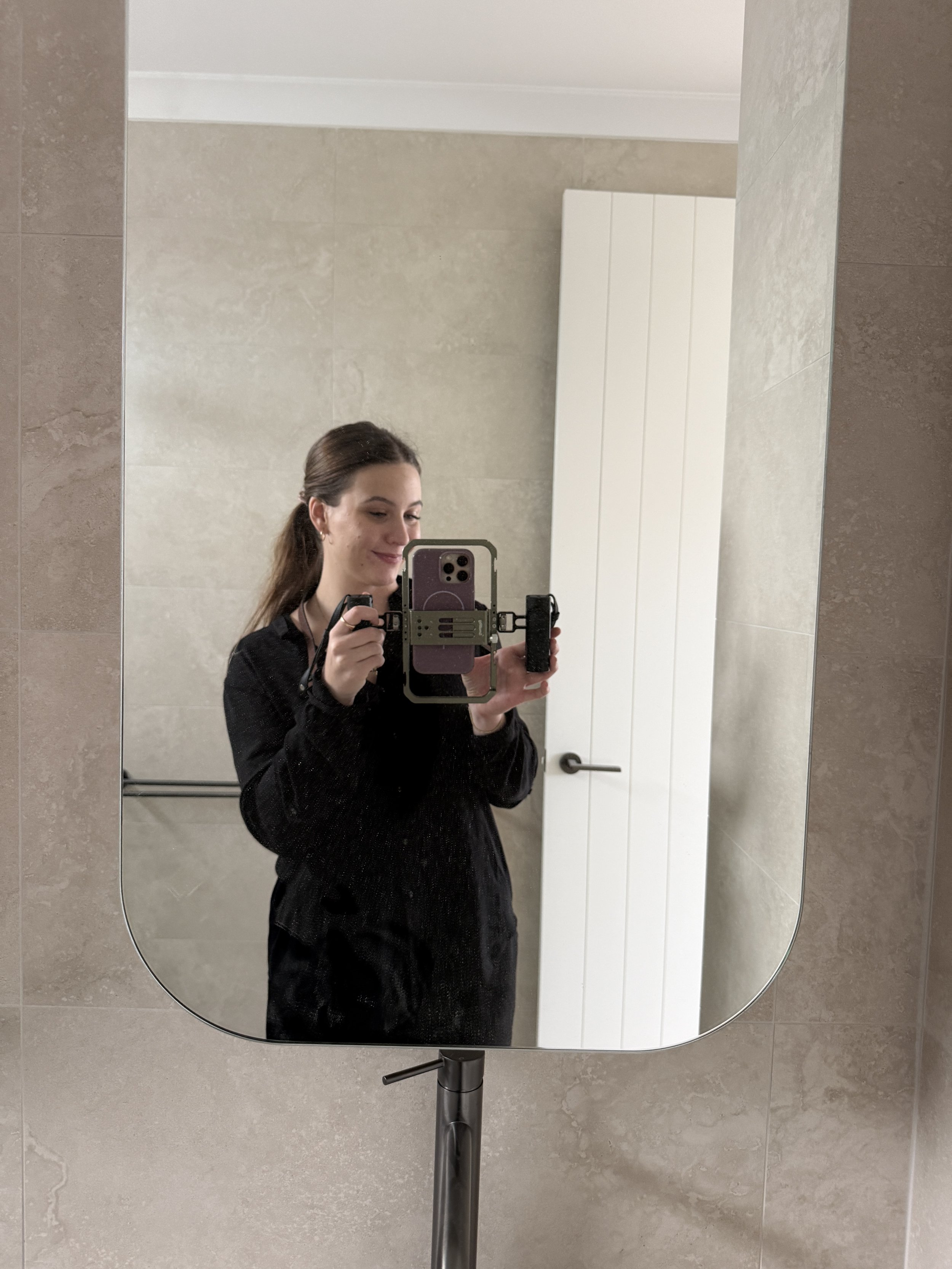 A young woman with brown hair tied back, dressed in black, taking a mirror selfie at a bathroom with beige tiled walls, holding a phone with a black case in one hand and a camera in the other.
