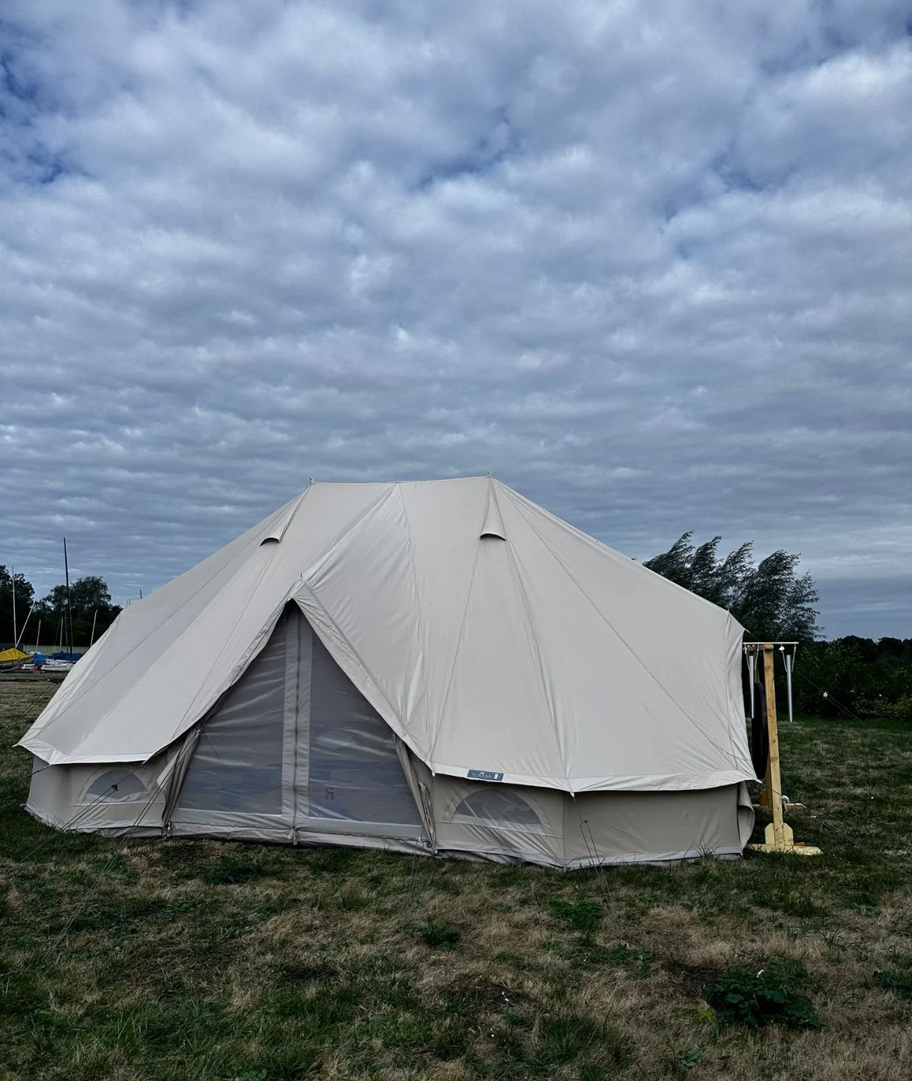 A large beige tent set up on a grassy field with trees and a cloudy sky in the background.