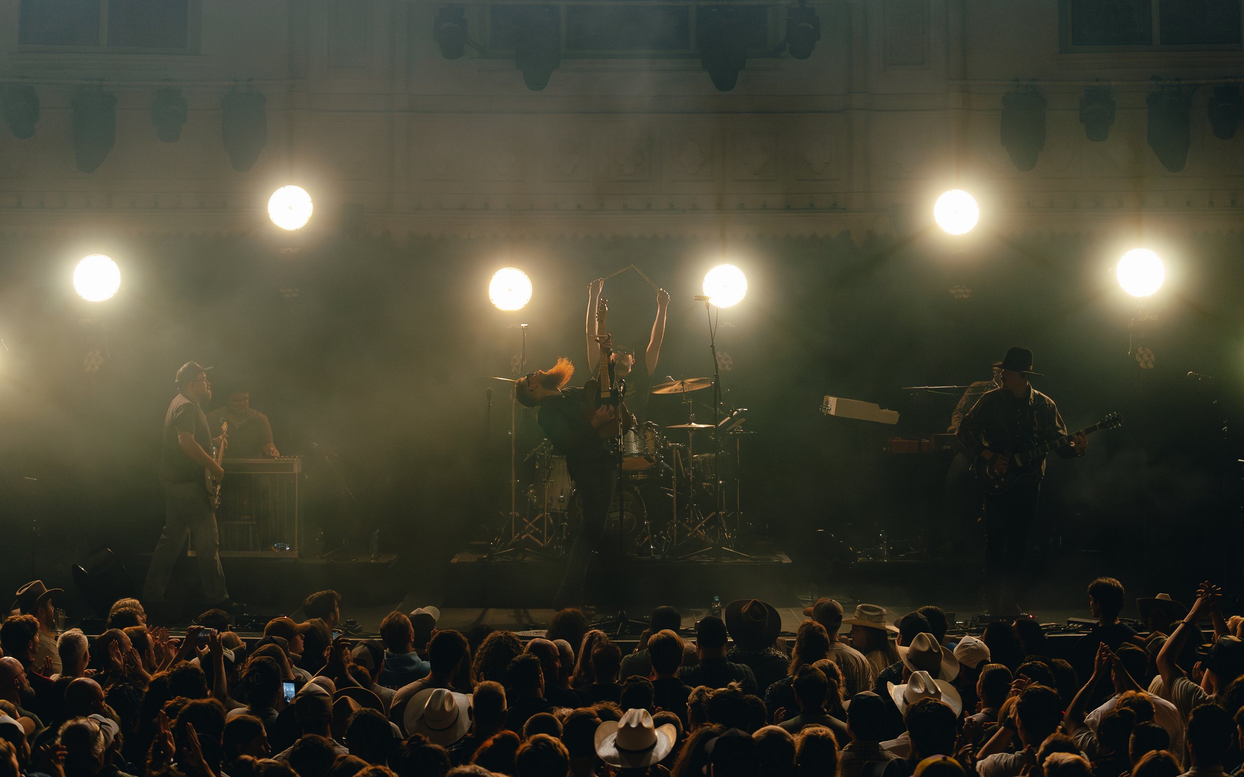 Live musical performance on stage with band members playing instruments, bright stage lights, and an audience wearing hats.