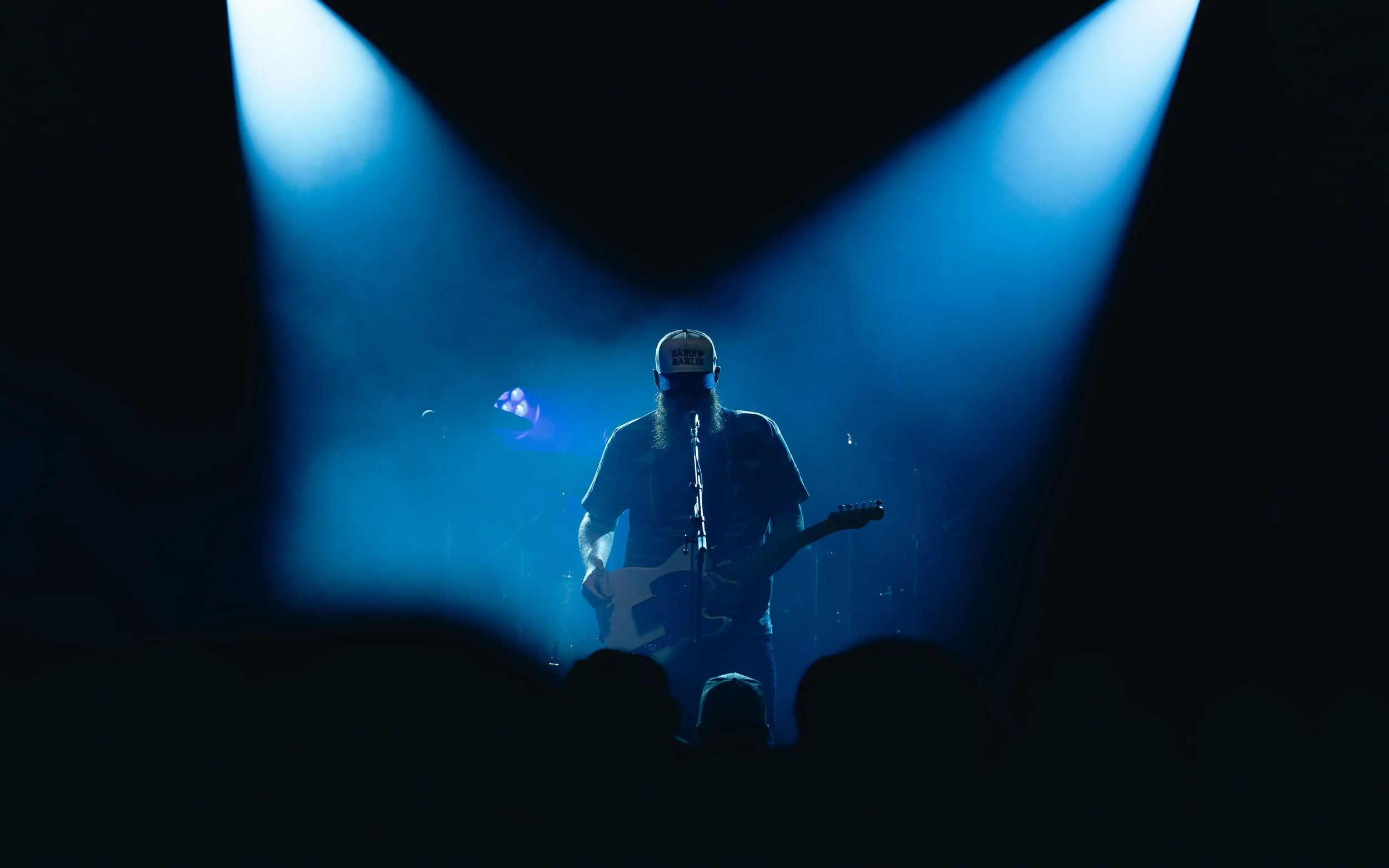 A musician playing an electric guitar on stage, illuminated by blue spotlights, with audience silhouettes in foreground.