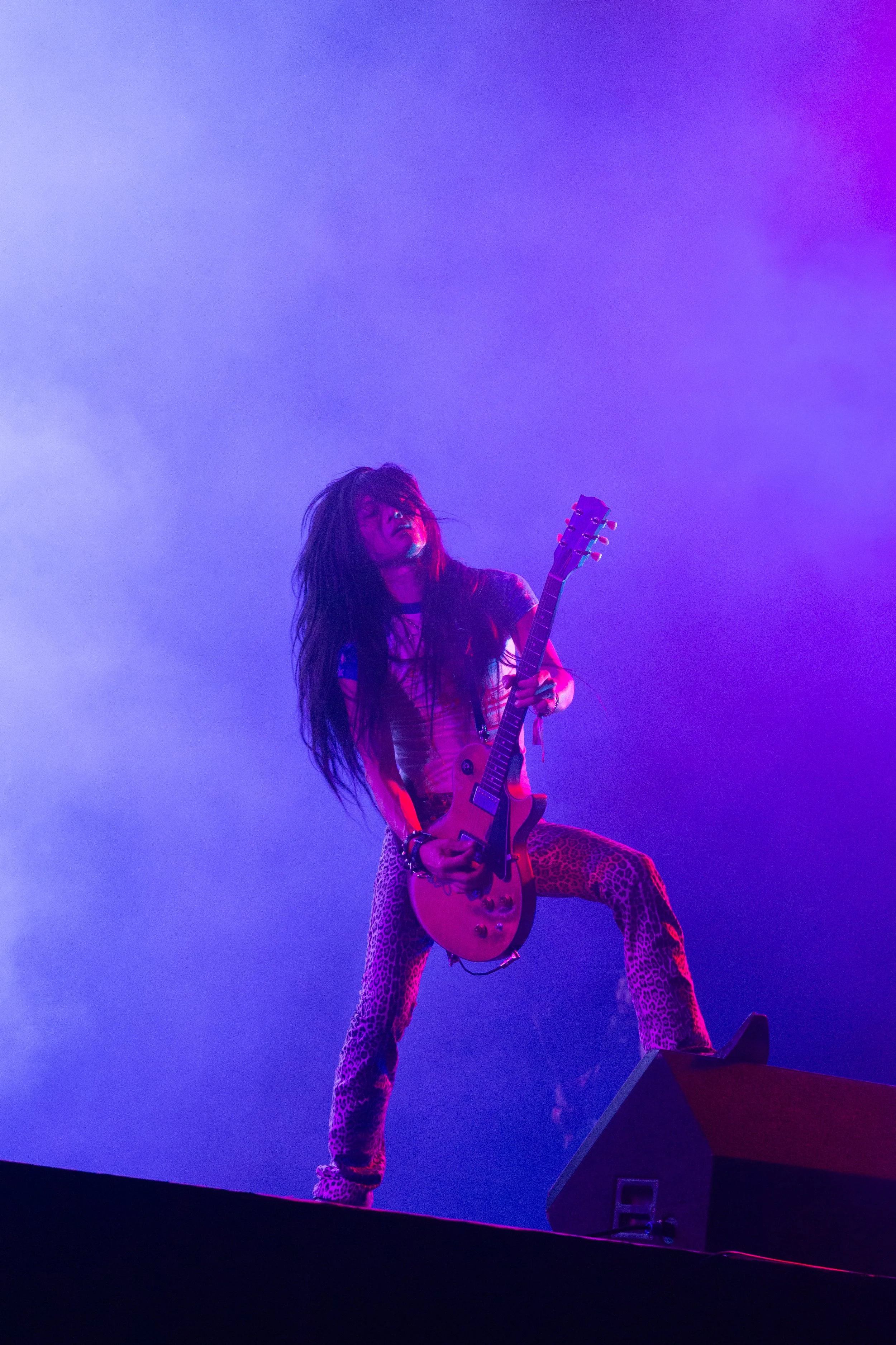 A female rock guitarist performs on stage, wearing leopard print pants and a colorful top, with purple stage lighting and a foggy atmosphere.