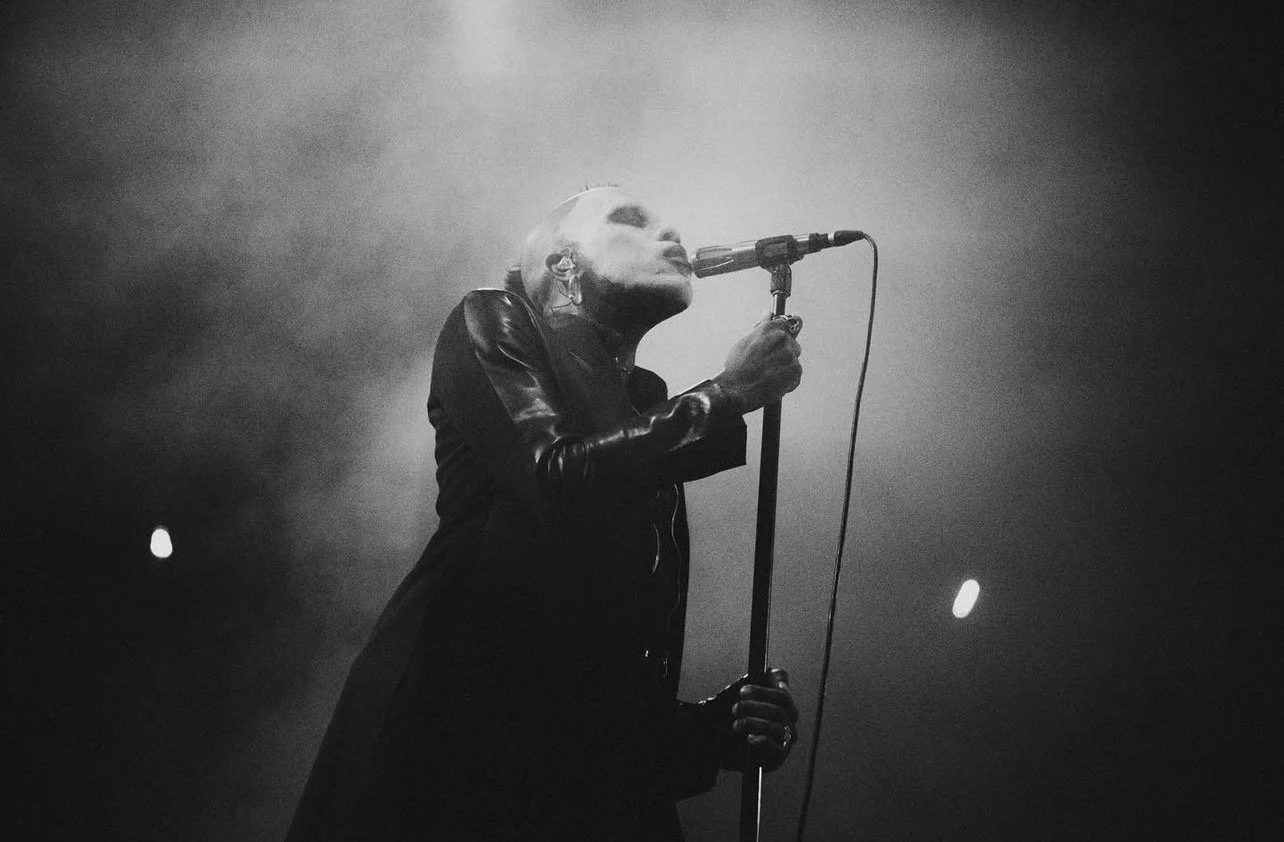 A black and white photo of a female singer passionately performing into a microphone on stage, wearing a leather jacket, with long hair, surrounded by fog or smoke.