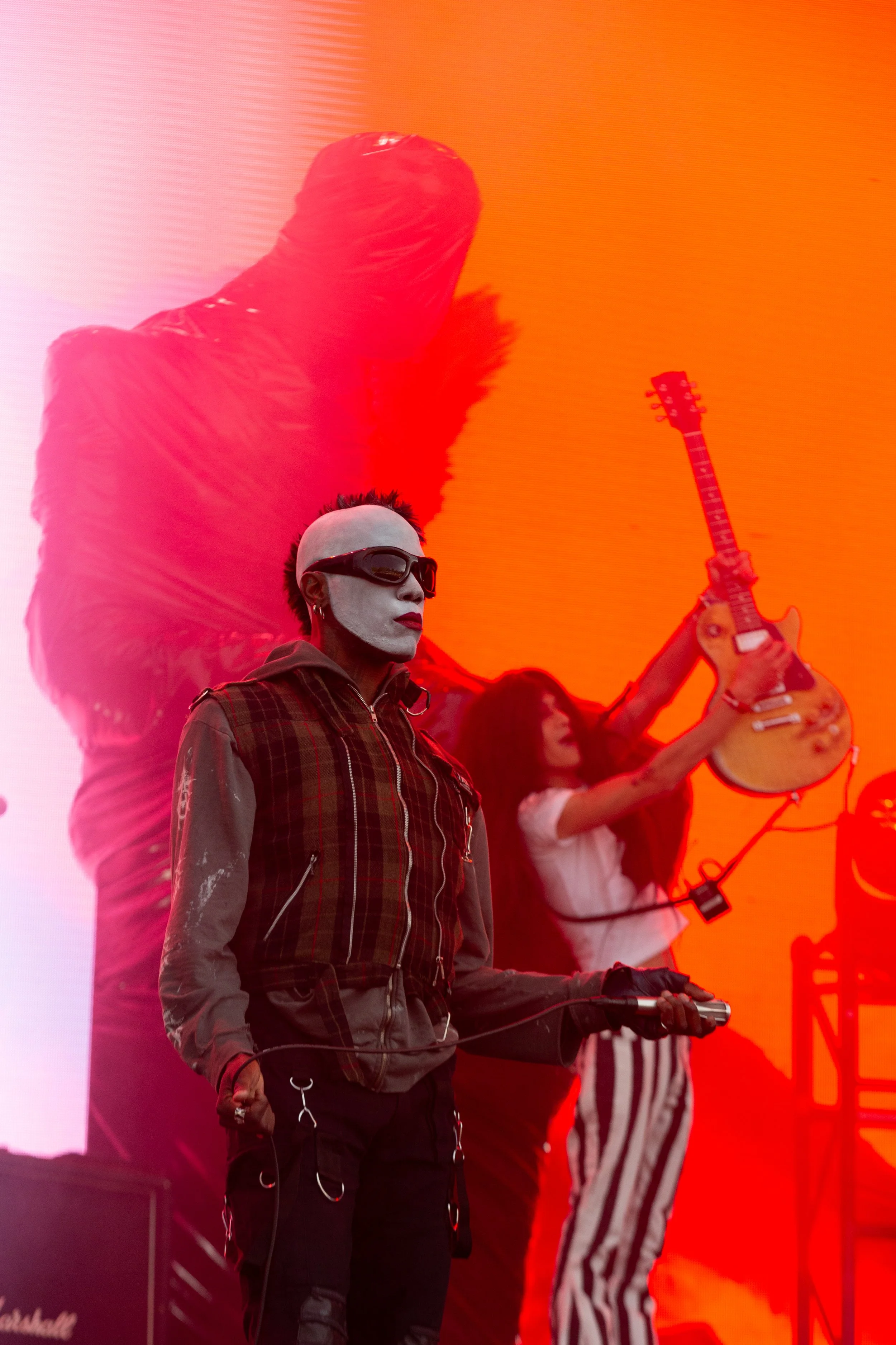 A performer with white face paint, black sunglasses, and spiked hair, holding a wireless microphone, stands on stage with a woman in the background playing an acoustic guitar, all against an orange backdrop with a silhouette of a large figure in the 
