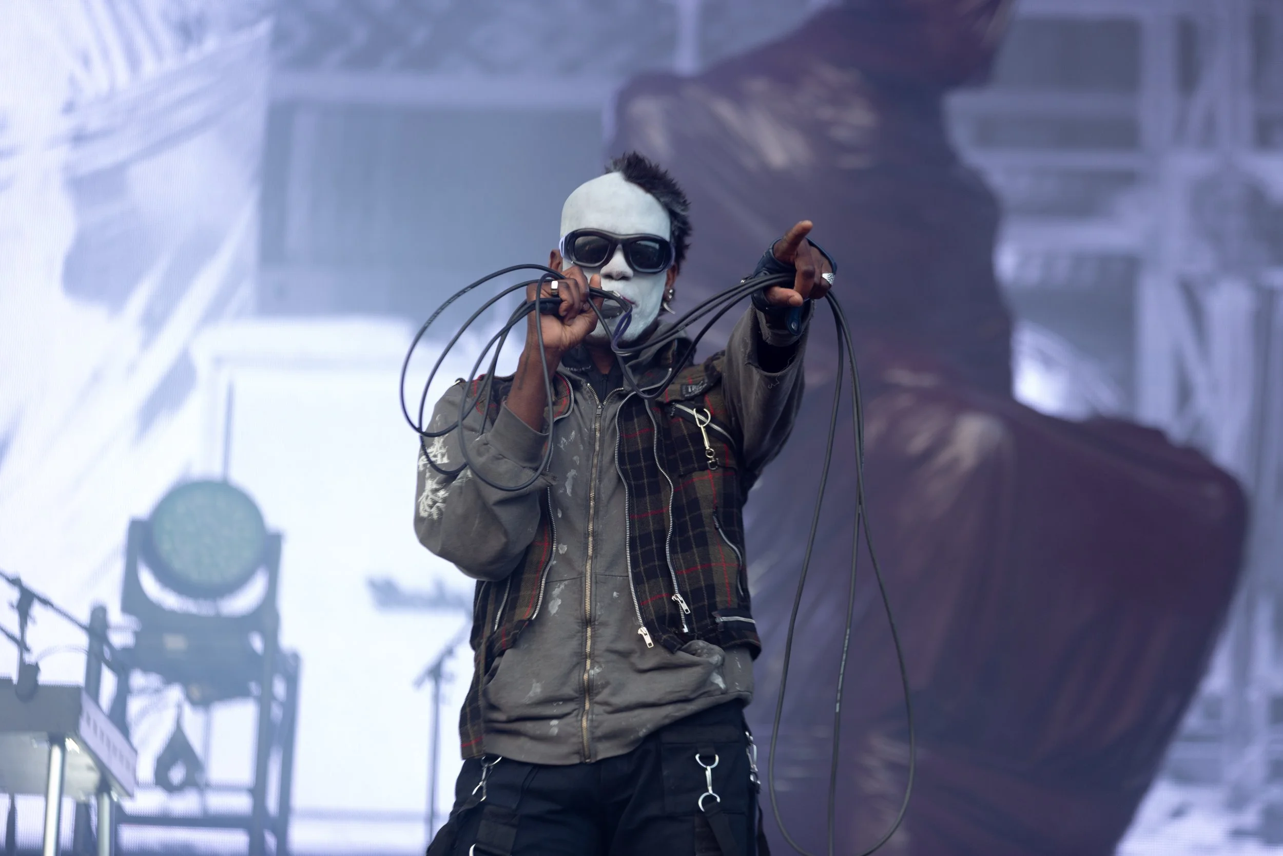 A performer on stage with white face paint, sunglasses, and a mohawk hairstyle, holding and pointing with a microphone wire, wearing a jacket and fingerless gloves.