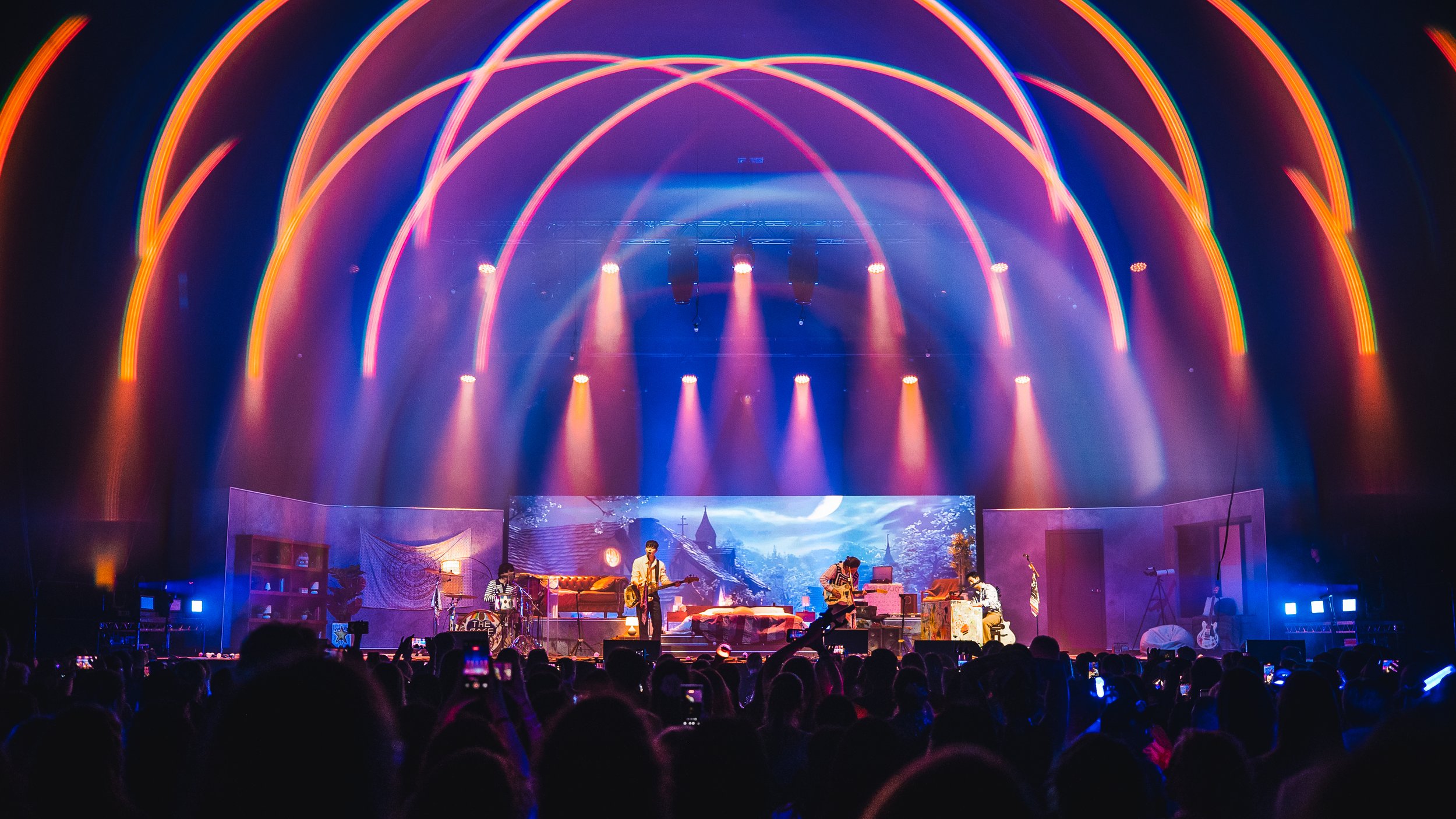 A concert stage with musicians performing, colorful neon lights above, and a crowd watching.