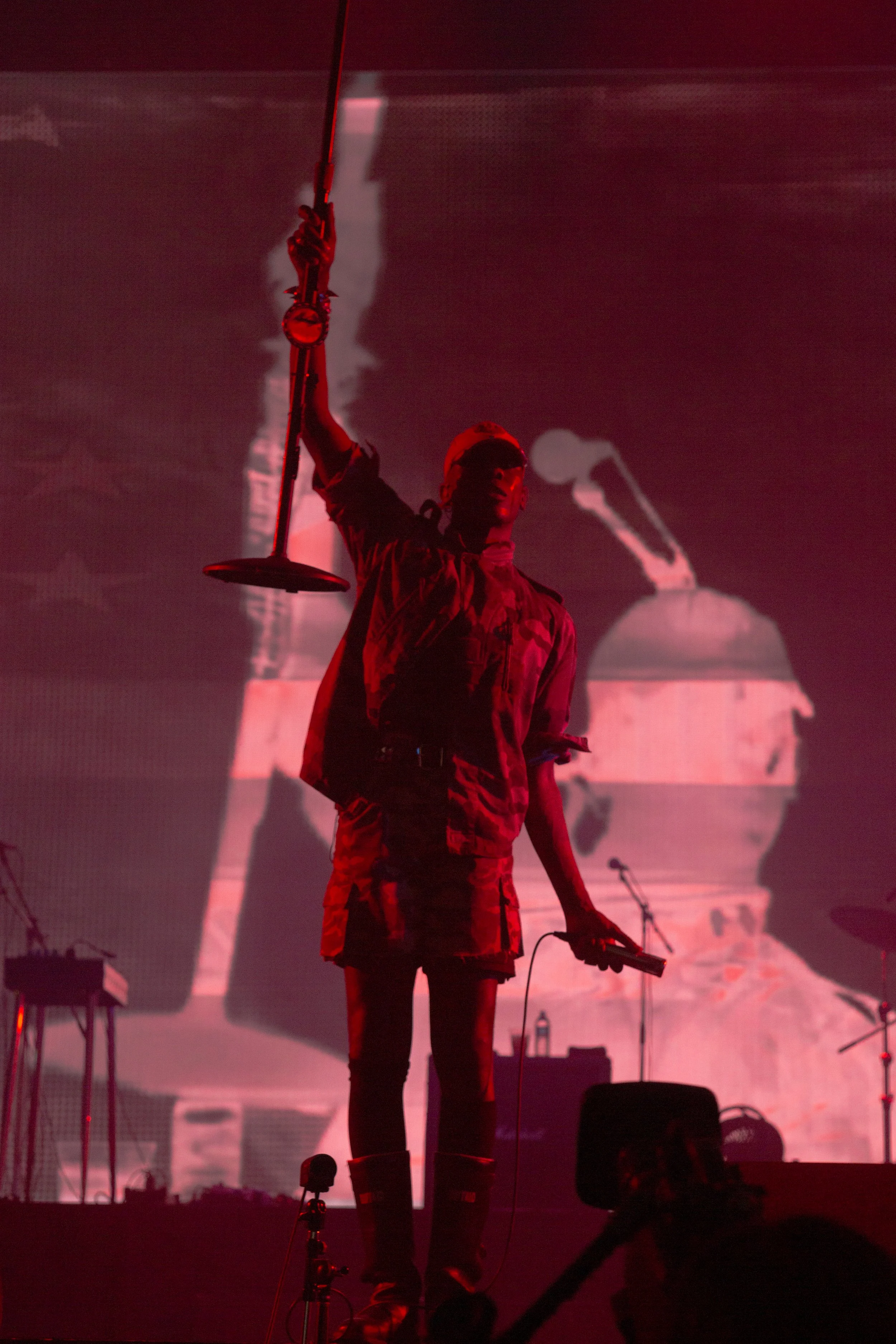 Person on stage holding a microphone stand, with a large screen in the background displaying a microphone and an object resembling a container or drum.