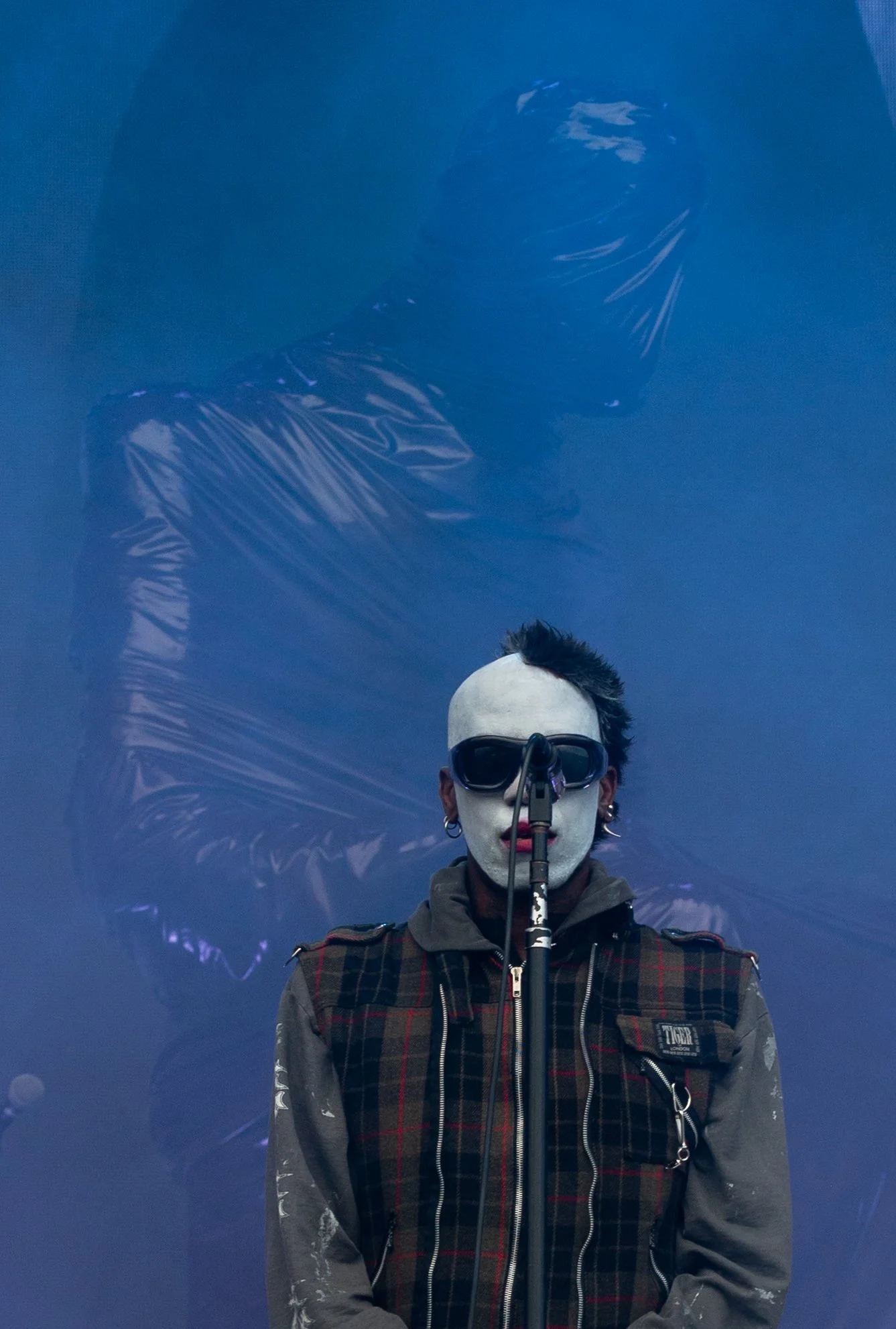 A person with white face makeup, black sunglasses, and a mohawk hairstyle, standing in front of a blue background with a large mirrored surface reflecting their image.