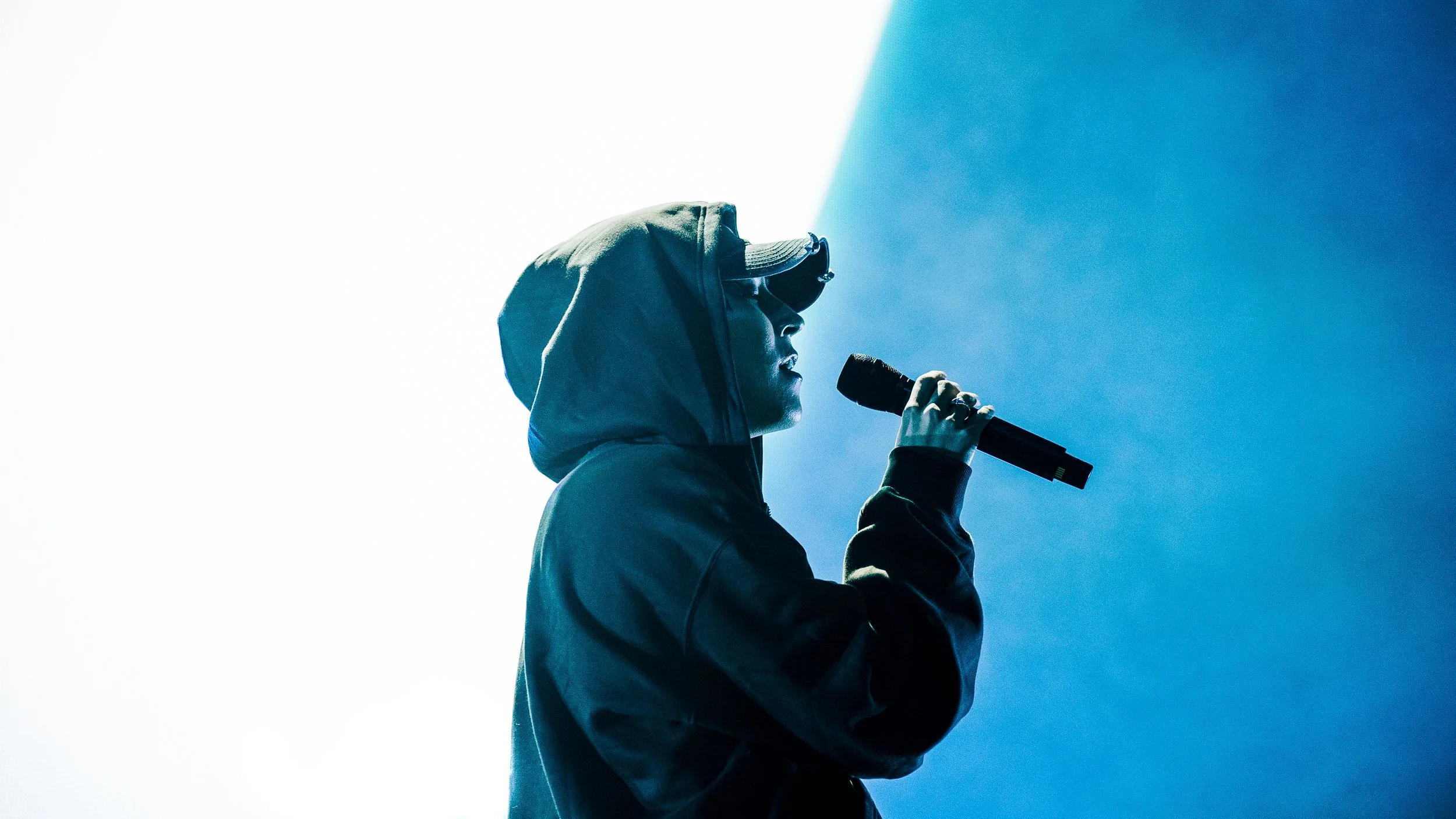 A person wearing a hoodie, sunglasses, and a baseball cap singing into a microphone on stage under blue lighting.