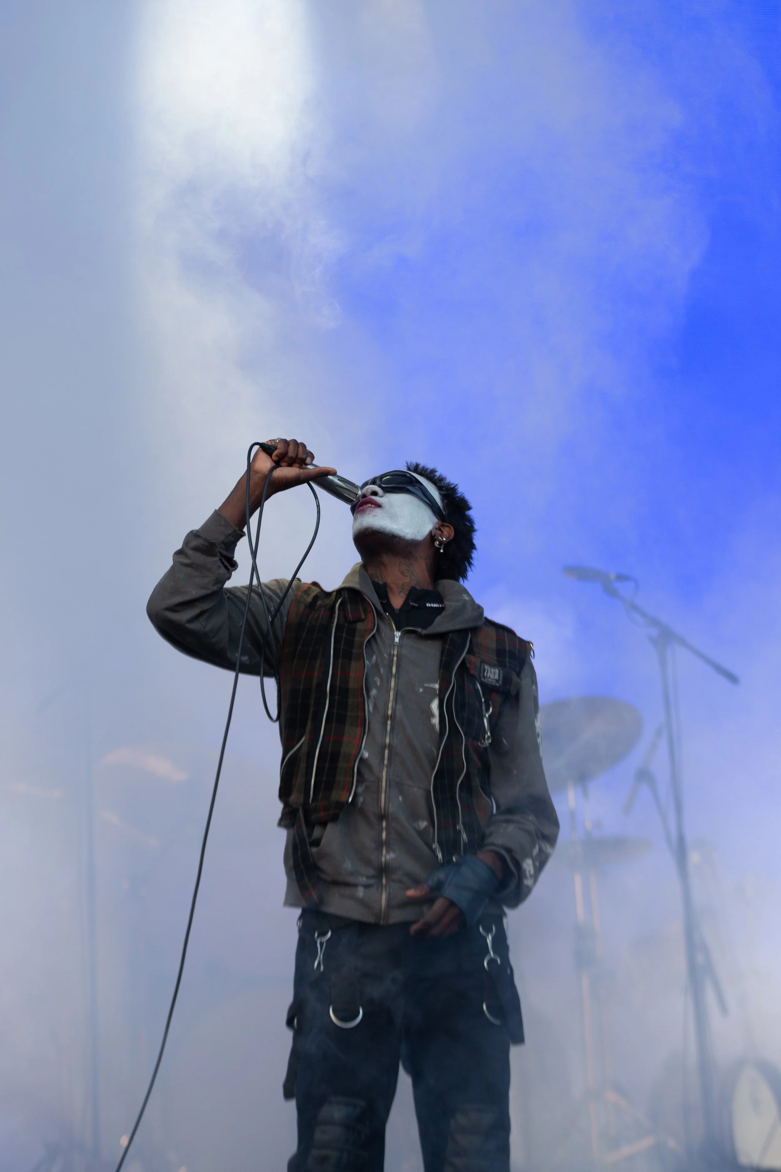 A male singer with face paint and sunglasses singing into a microphone on stage with a blue sky and clouds in the background.