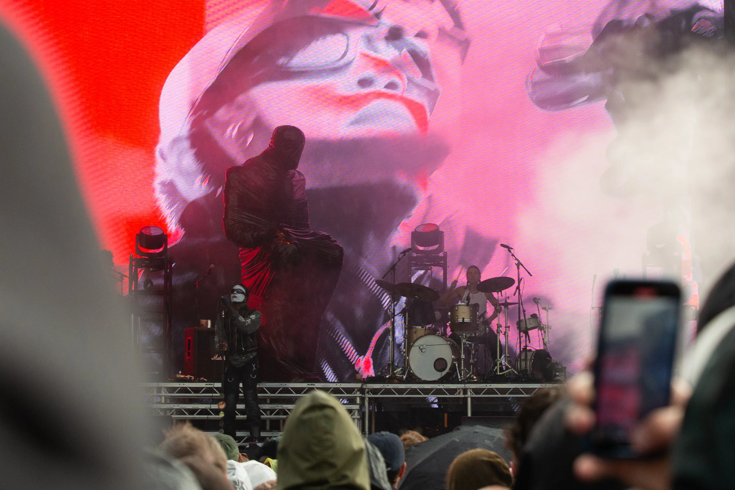 A live concert with a drummer, a singer with face paint, and a large LED screen showing an image of a woman with white glasses and red makeup. The audience is visible in the foreground, some recording with phones.