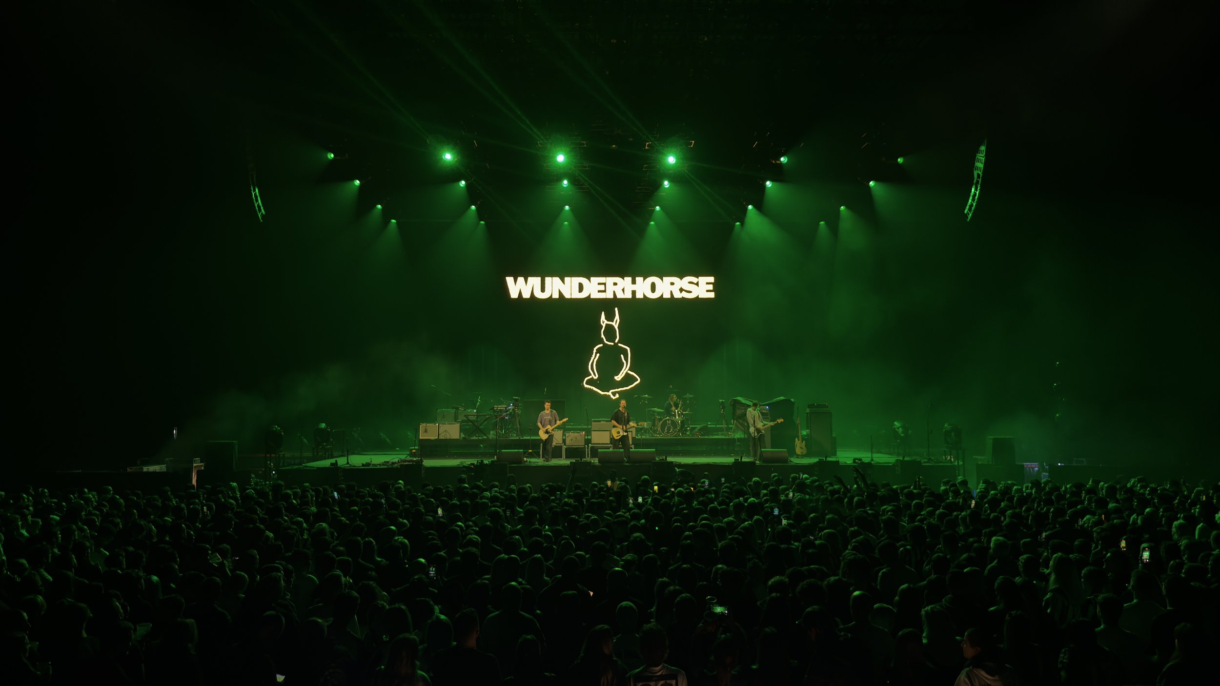 A concert stage illuminated with green lights with musicians performing and a large screen displaying the word 'WUNDERHORSE' above a bunny logo.