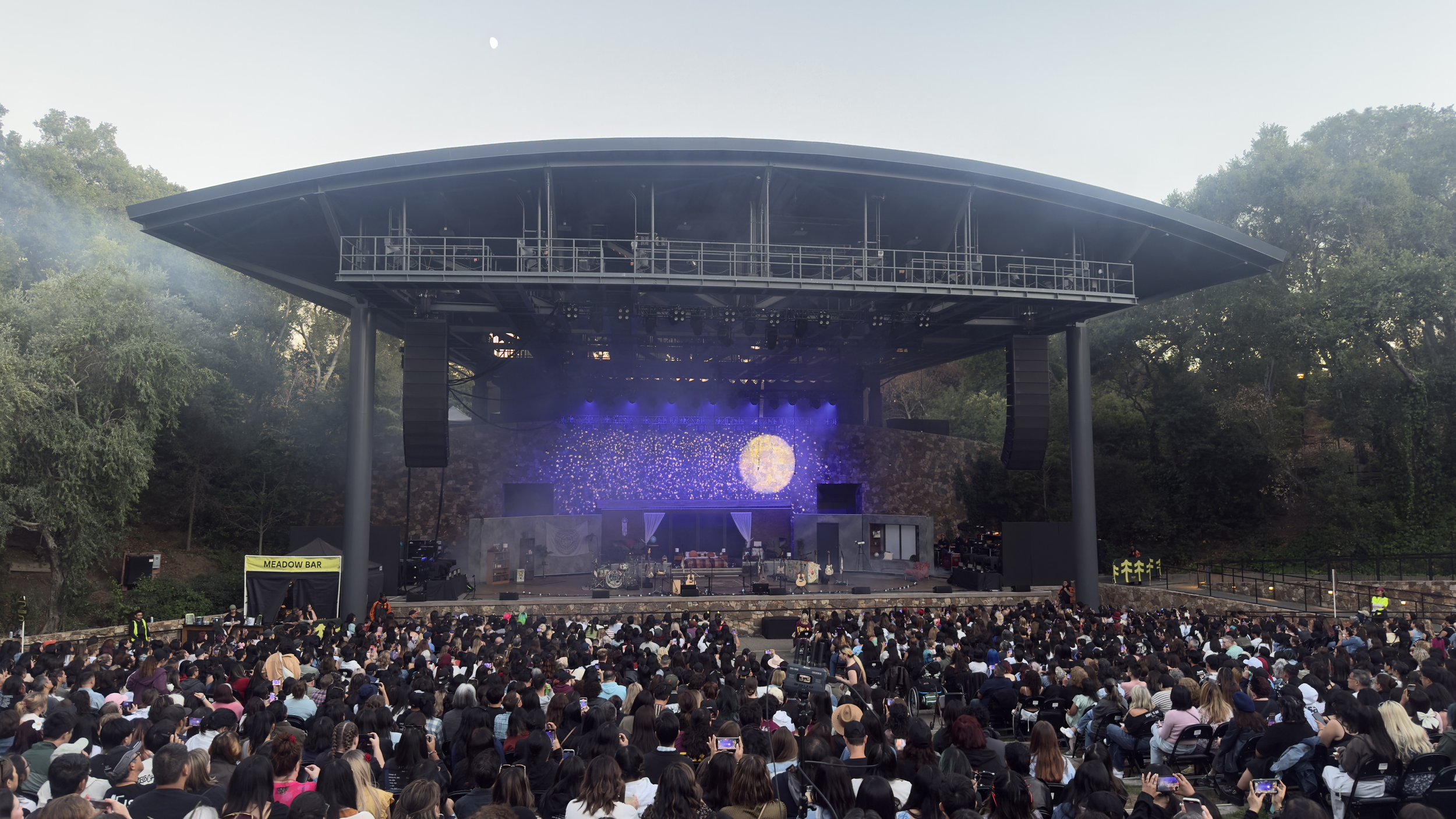 An outdoor concert with a large crowd, a stage with musical instruments, and a screen displaying a starry night scene, surrounded by trees.