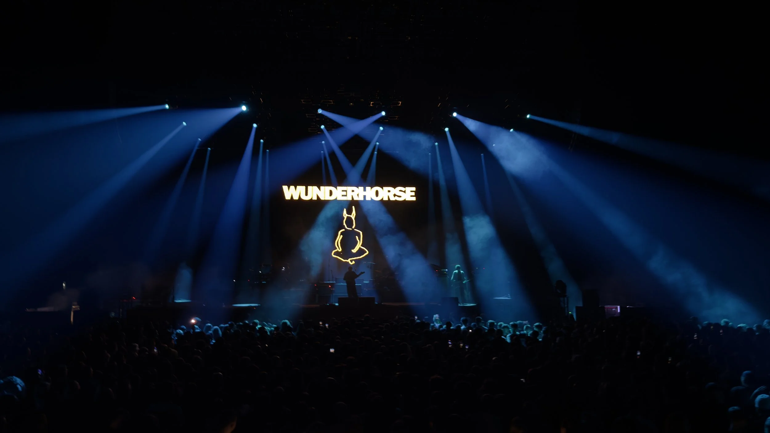 Concert scene with blue stage lights, a large screen displaying the text 'WUNDERHORSE' and a line drawing of a rabbit sitting cross-legged, with a crowd in the foreground.
