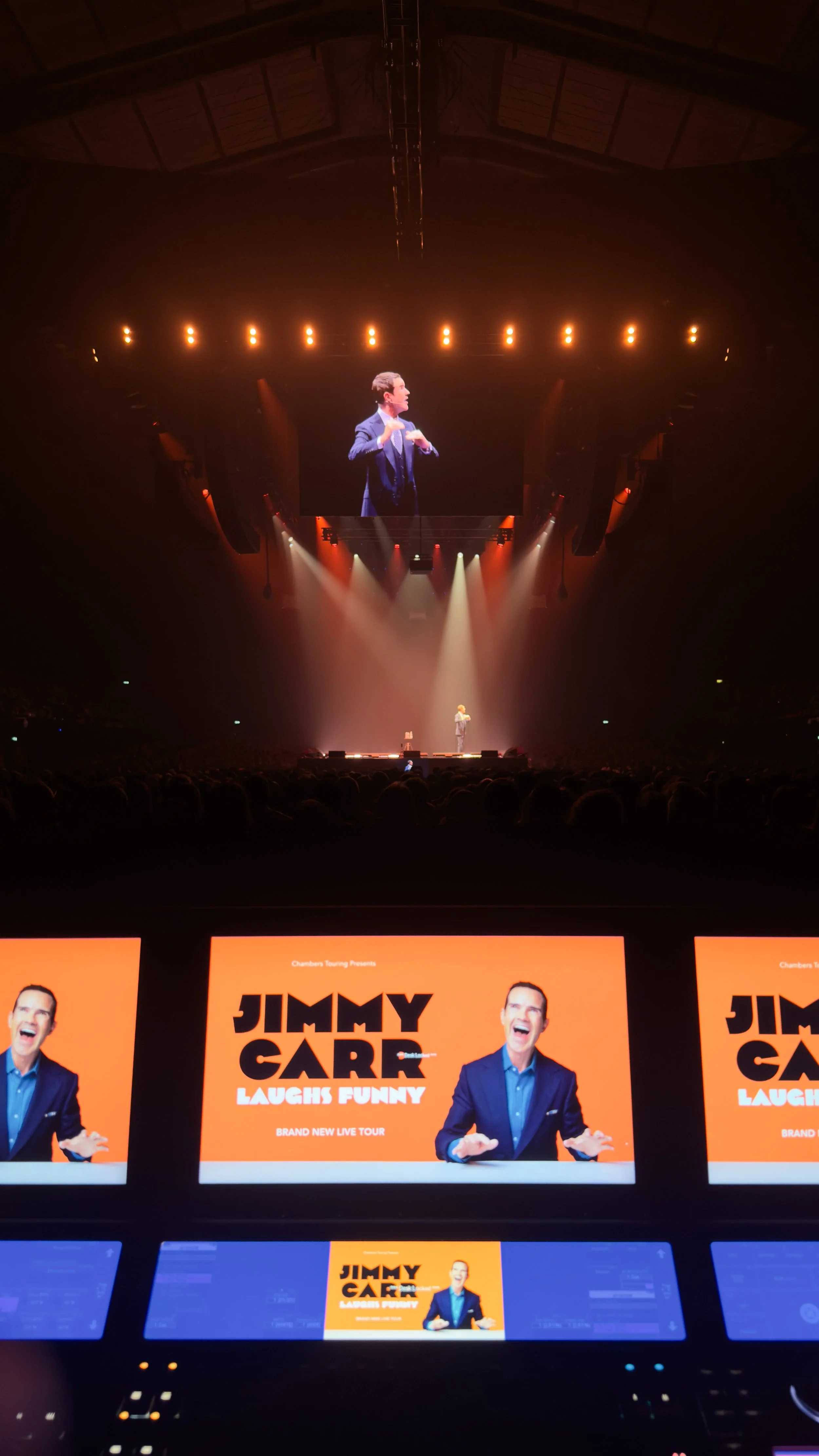 A stage with a large screen showing a man in a suit, and several smaller screens displaying promotional material for Jimmy Carr's comedy tour, titled 'Laughs Funny'.