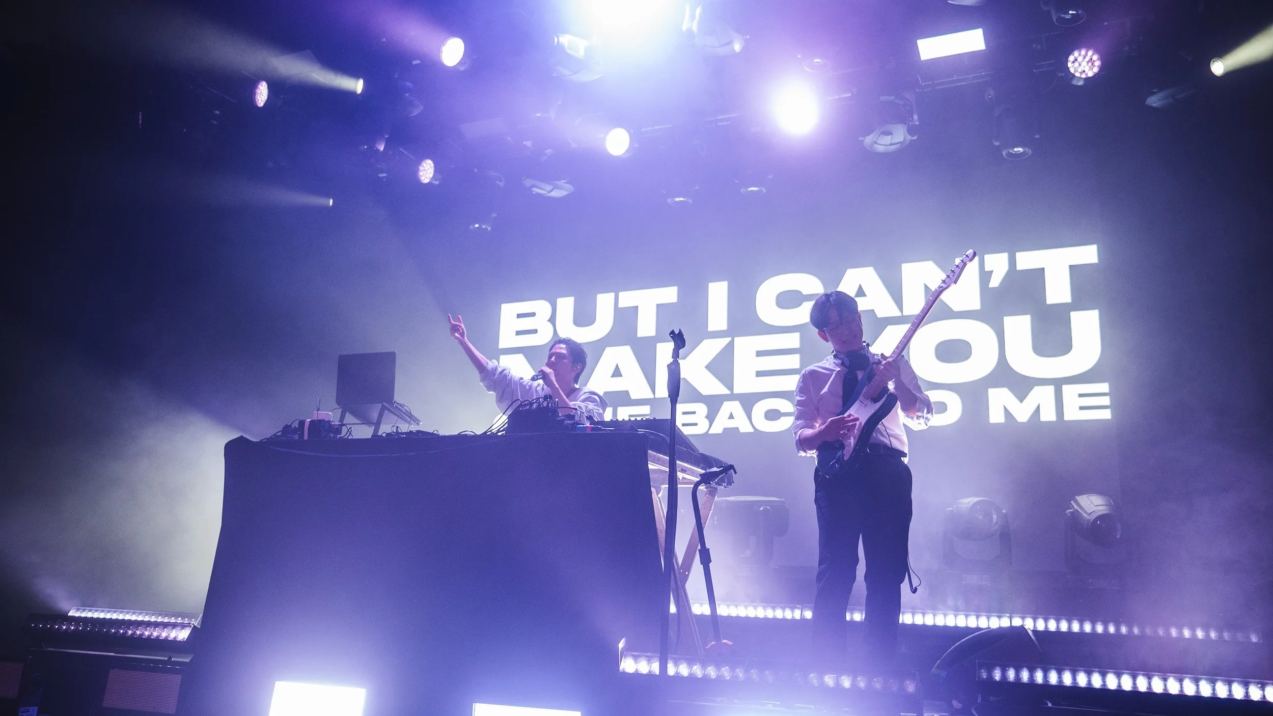 Two musicians on stage, one with DJ equipment and the other playing an electric guitar, with a large screen behind them displaying the lyrics 'BUT I CAN'T MAKE YOU REMEMBER ME' in white text.