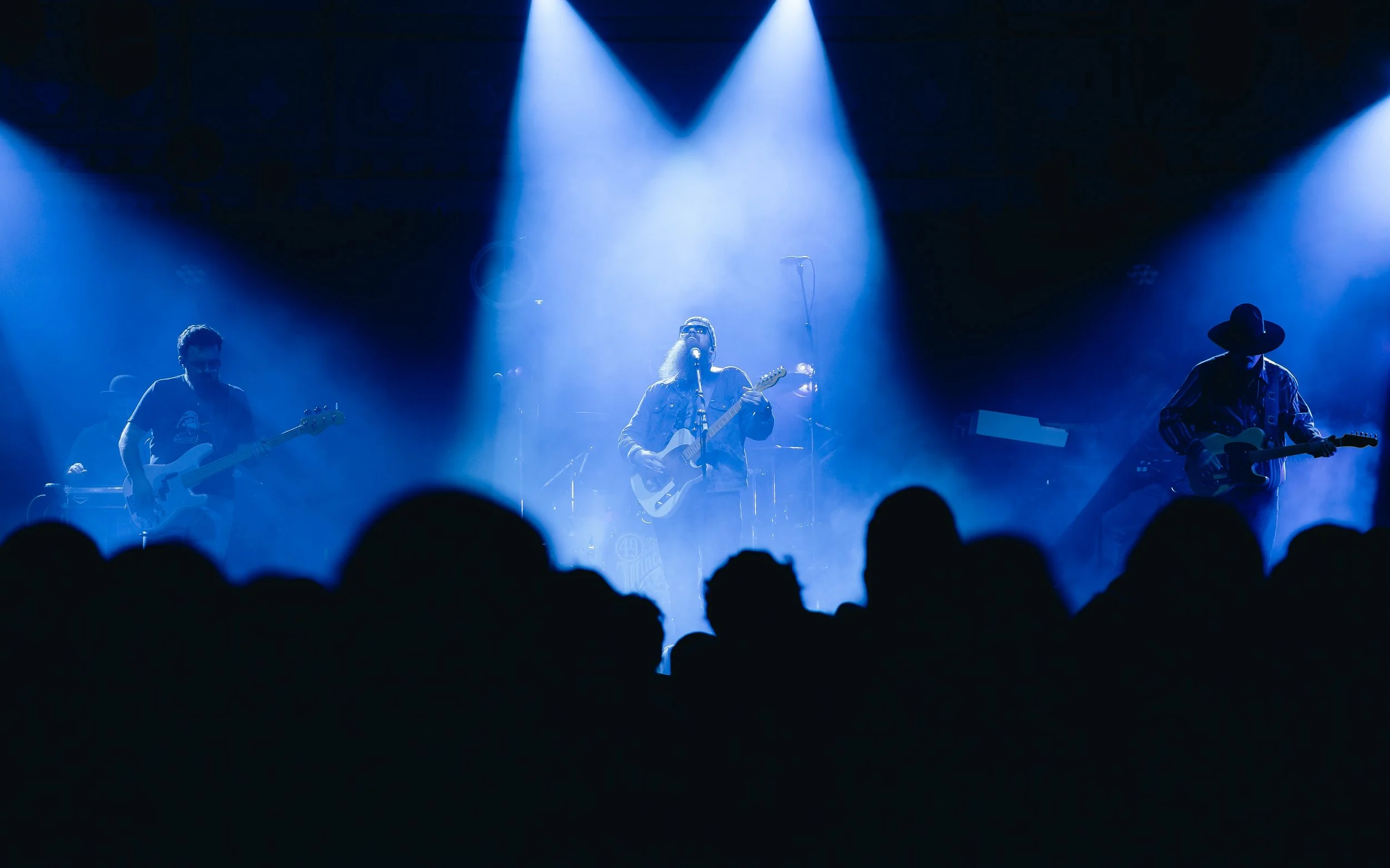 A band performs on stage with blue lighting, including a singer with glasses and a beard, a guitarist with a hat, and a bassist, with an audience visible in the foreground.