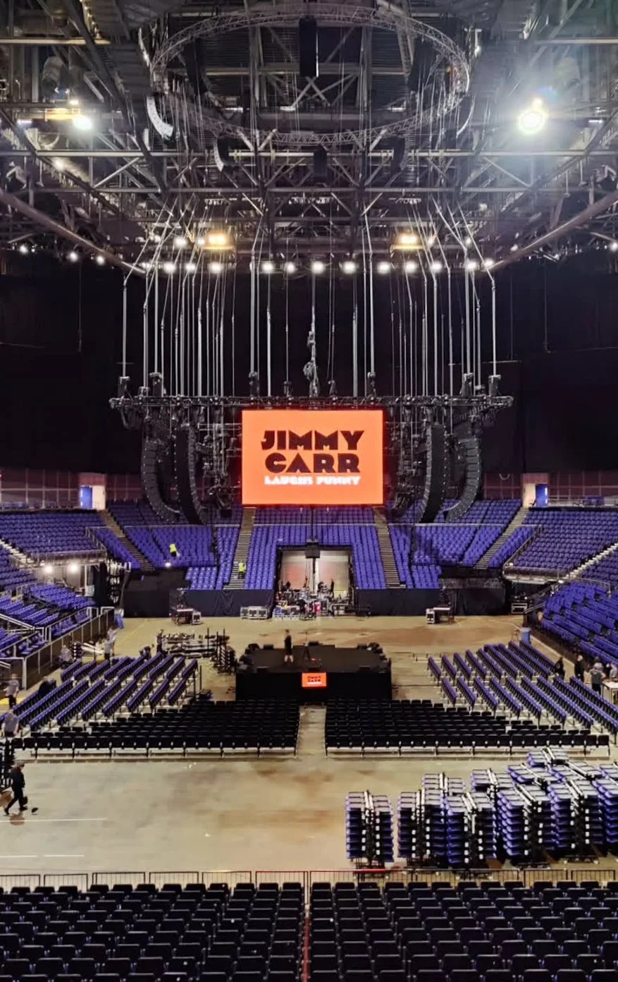 Concert stage setup in an arena with a large screen displaying Jimmy Carr's name, surrounded by stage rigging and empty seating.