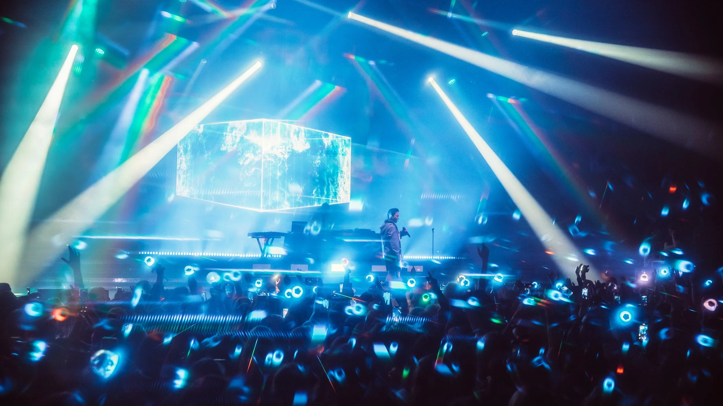A DJ performs at a concert with vibrant blue and white lights, a large screen displaying visuals, and an enthusiastic crowd holding glowing accessories.