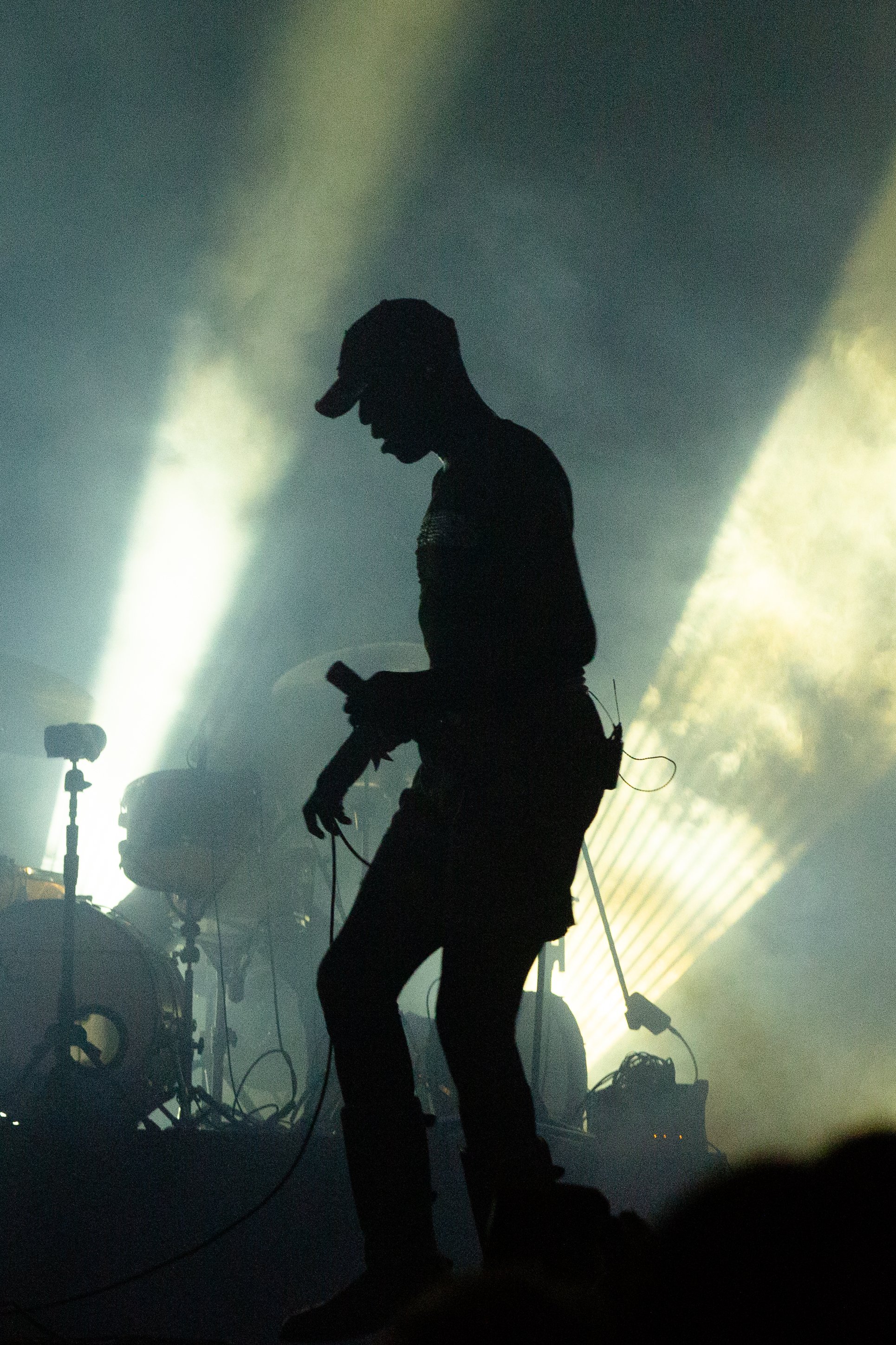 Silhouette of a person wearing a cap, holding a device, possibly a microphone or remote, standing on stage with bright lights and fog in the background.