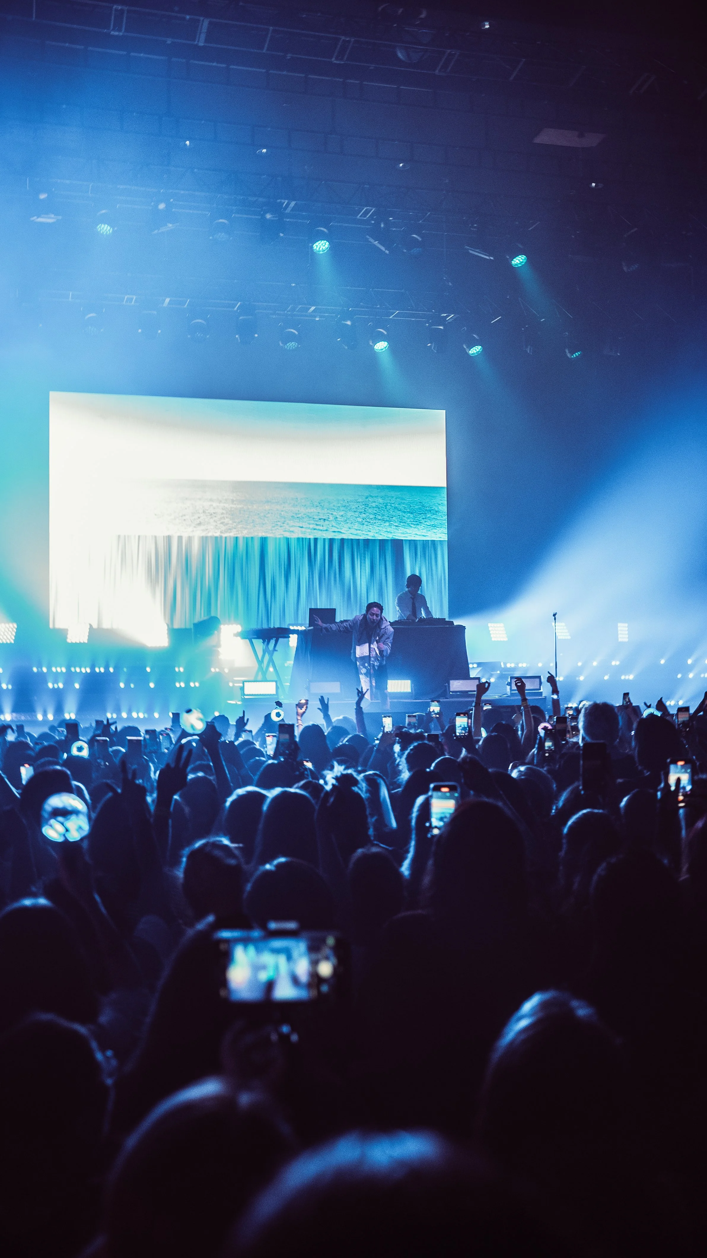 A concert scene with a large crowd, a performer on stage, bright blue and white stage lights, a large digital screen displaying an ocean scene, and audience members capturing the moment on their smartphones.