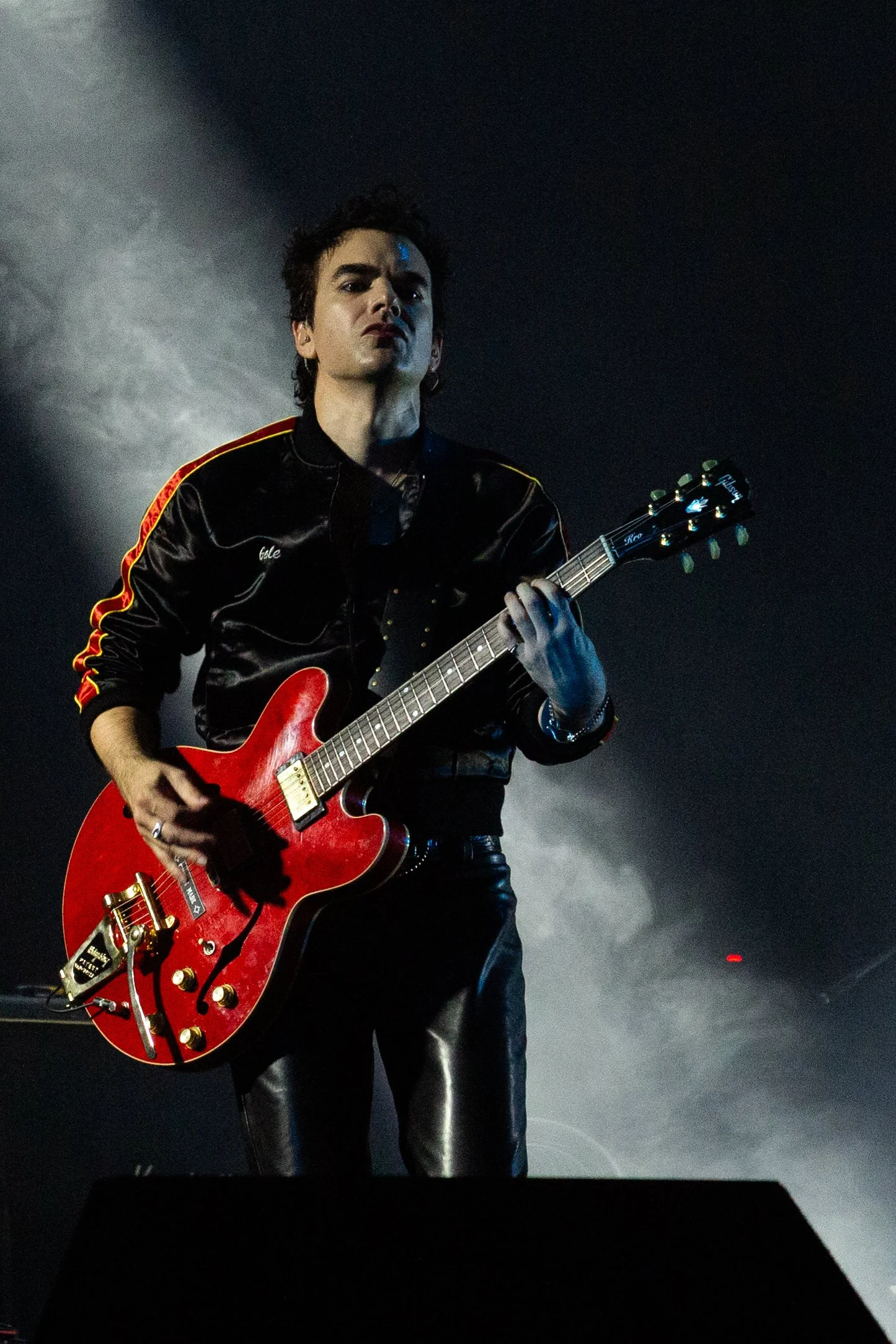 A male guitarist performs on stage with a red semi-hollow body electric guitar, wearing a black satin jacket with red and yellow accents, and black leather pants, under dramatic lighting with smoke in the background.