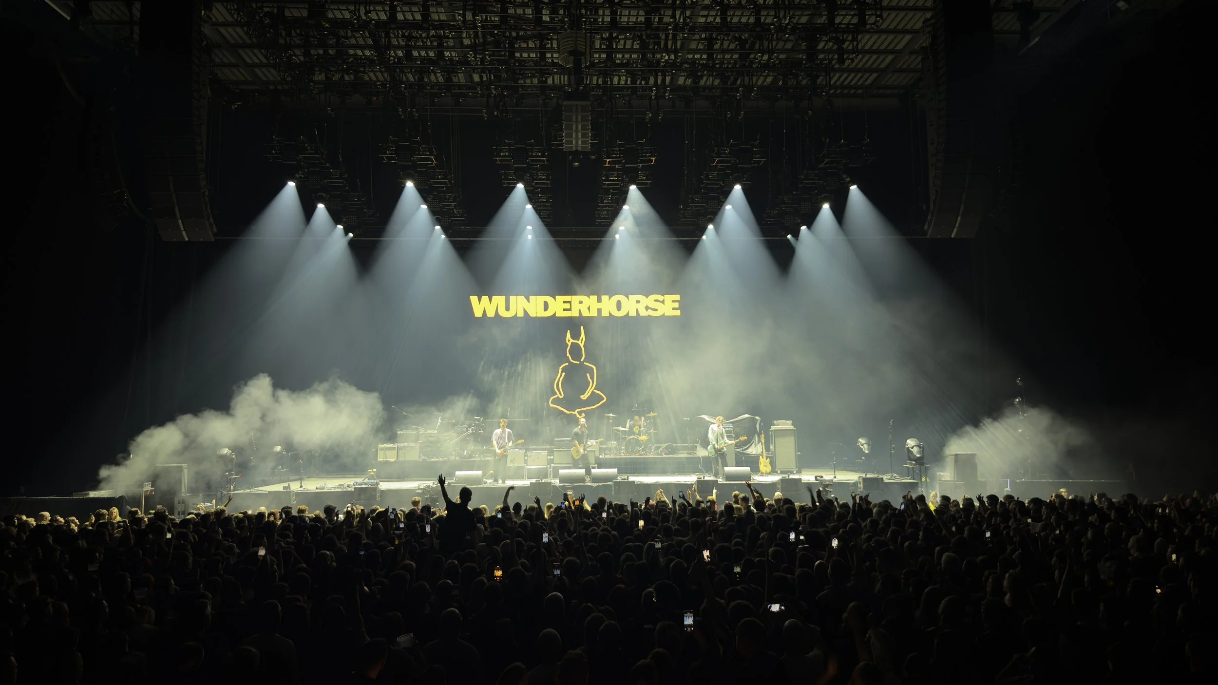 Concert stage with four musicians performing, illuminated by overhead spotlights, fans in the audience, with a large screen displaying the word 'WUNDERHORSE' and a rabbit-like figure in yellow outline.