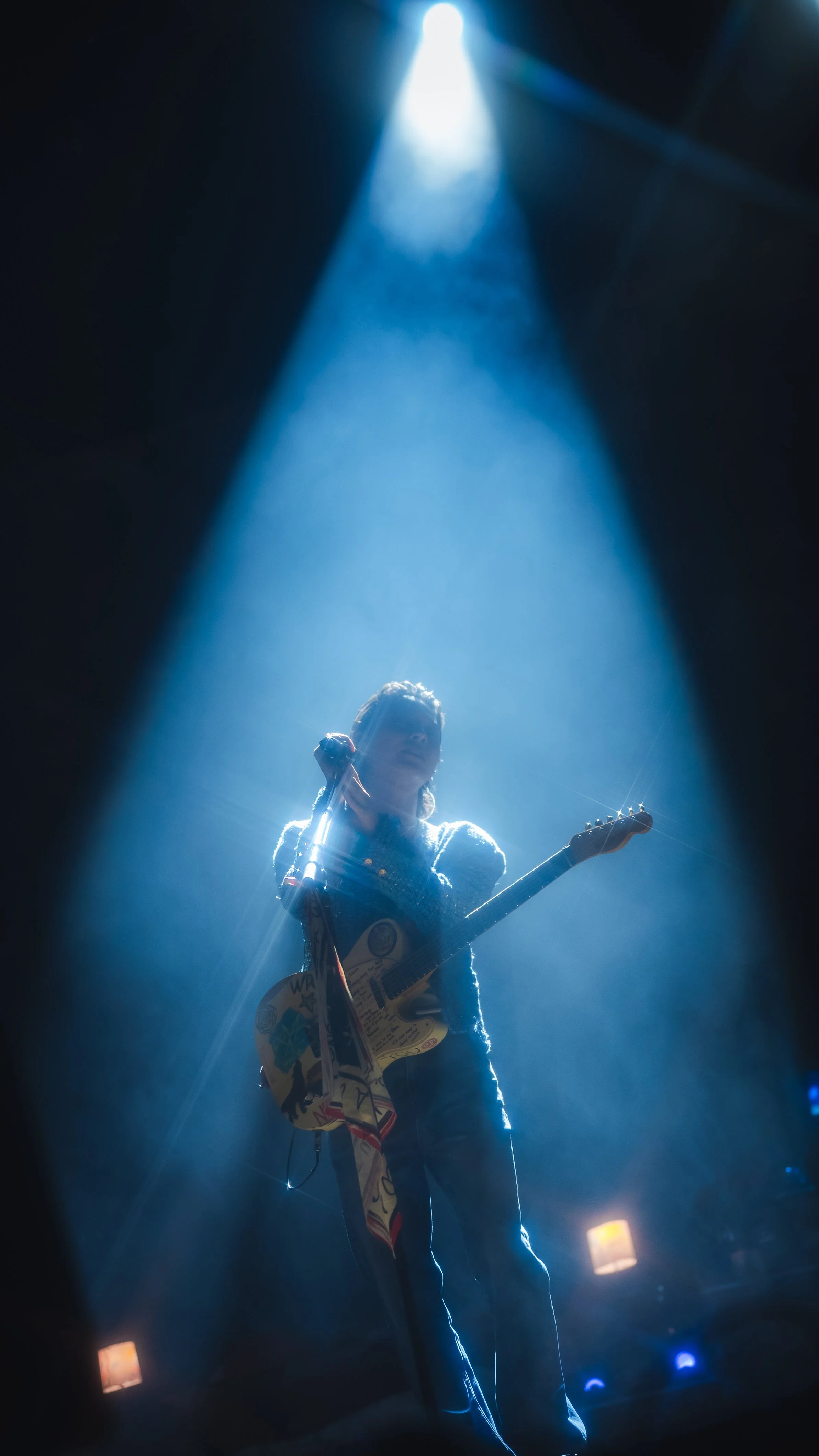 A musician on stage holding a guitar, illuminated by a spotlight in a dark setting, with smoke or fog creating a dreamy atmosphere.