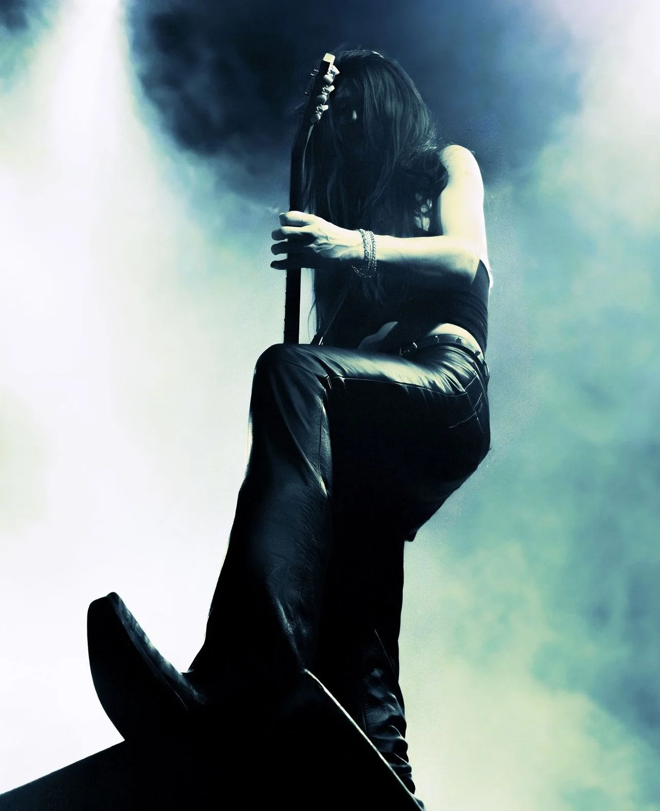 A female musician playing an electric guitar on stage, dressed in black leather pants and a sleeveless top, with long dark hair covering her face, in a dynamic performance with fog and dramatic lighting.