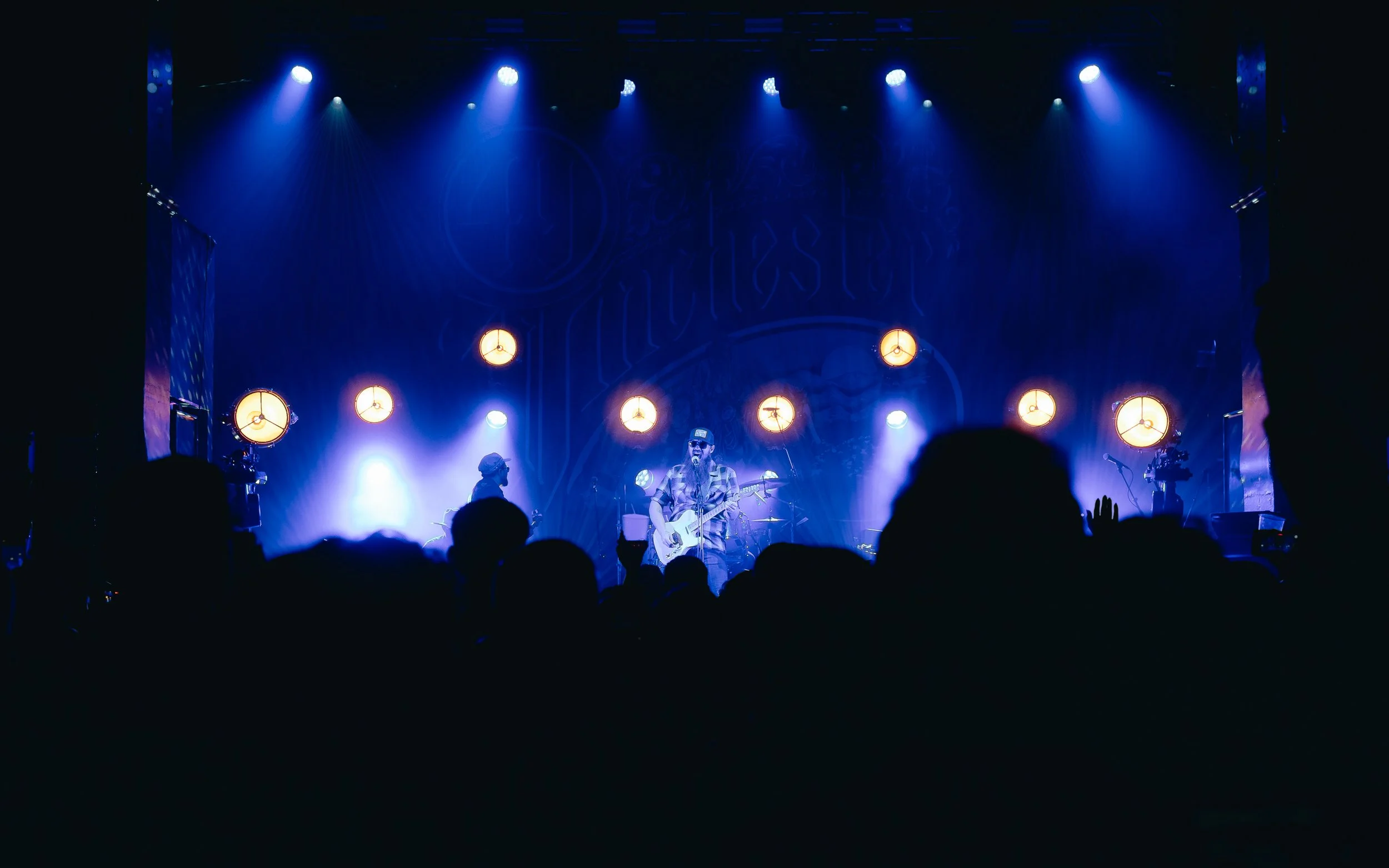 A live music concert with a band performing on stage and audience silhouettes in the foreground, illuminated by blue and yellow stage lights.