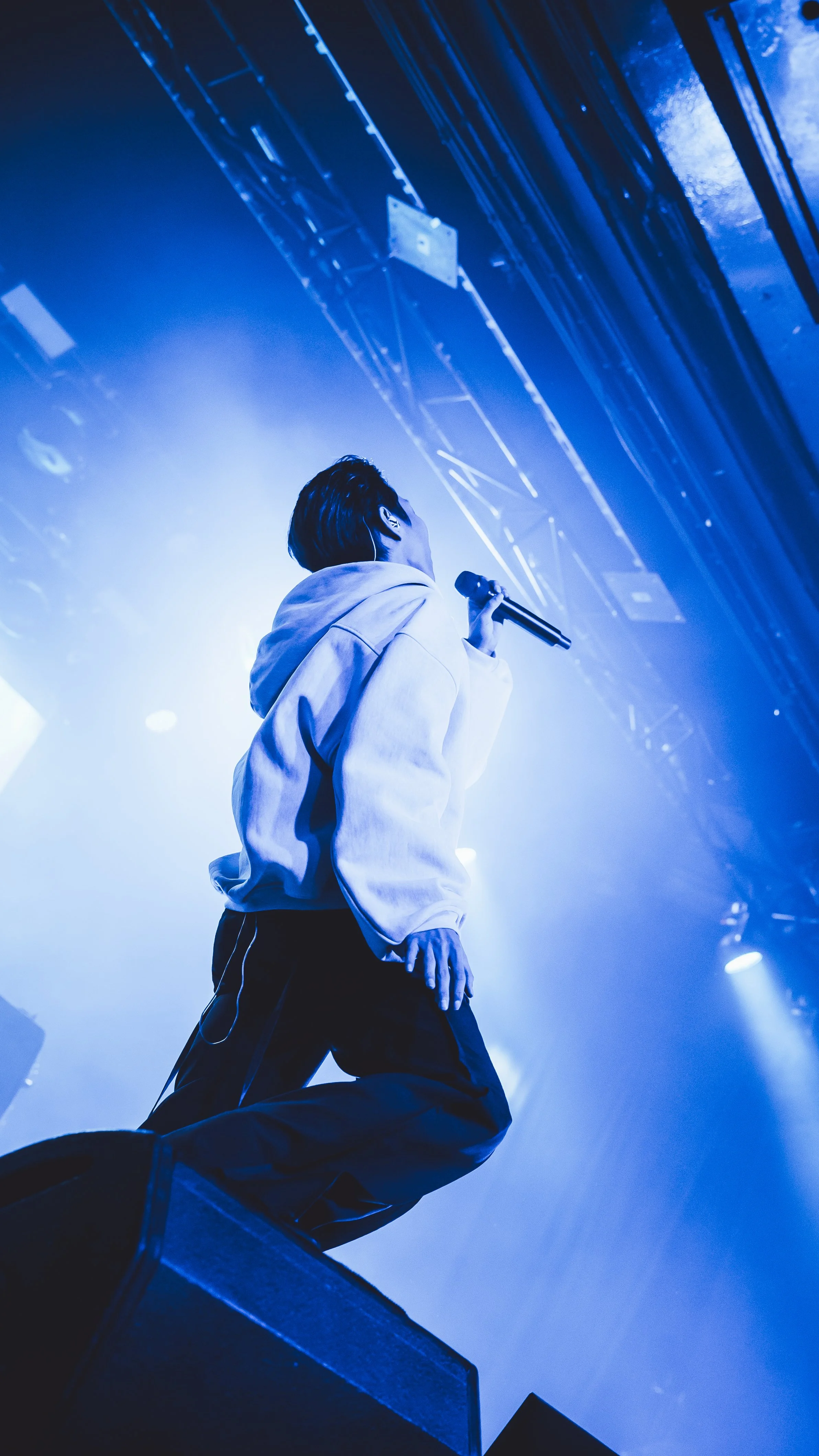 Performer on stage holding a microphone during a concert, illuminated by blue stage lighting, wearing a light-colored hoodie and dark pants, with stage scaffolding overhead.