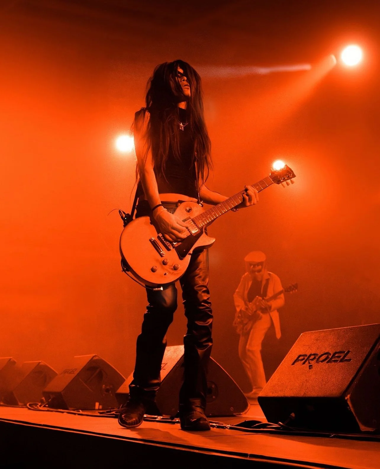 Male guitarist with long hair and dark clothing performing on stage, with orange lighting and a second guitarist in the background.