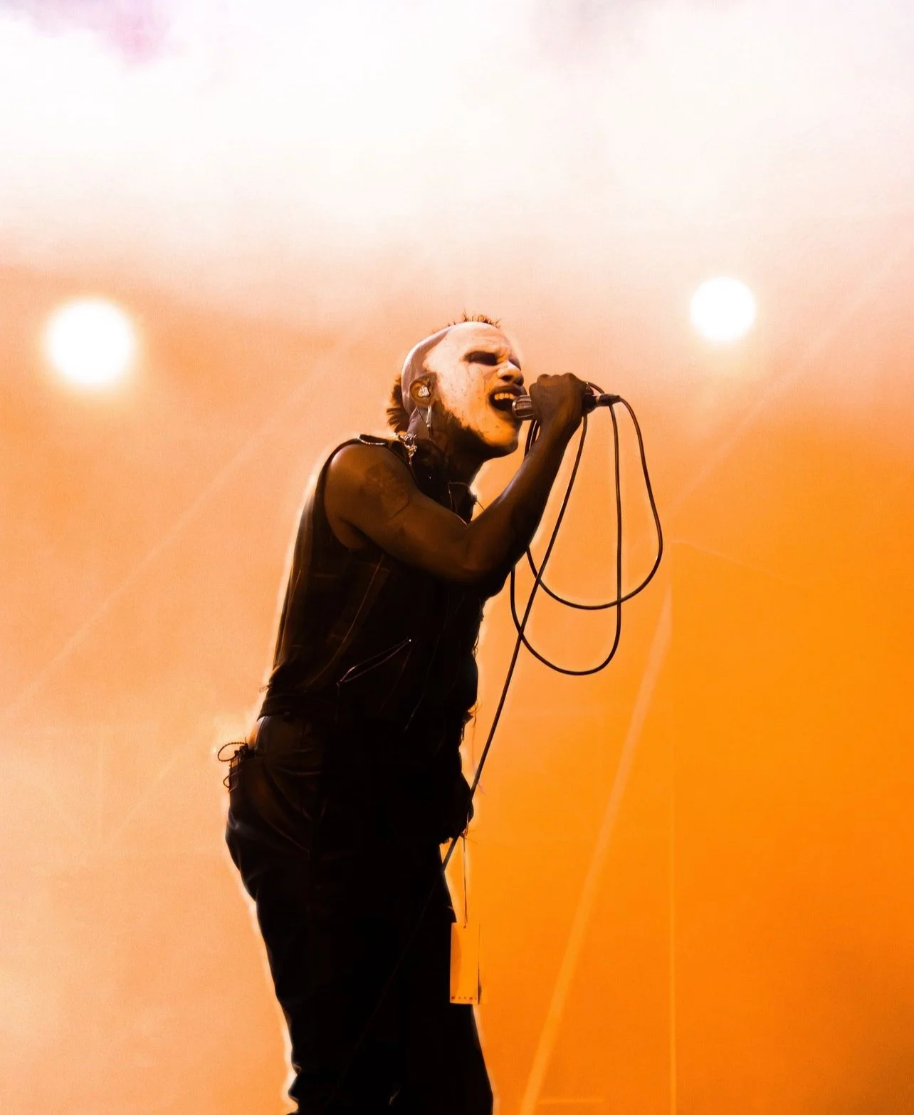 A male singer with spiked hair and tattoos singing passionately into a microphone on stage with orange lighting