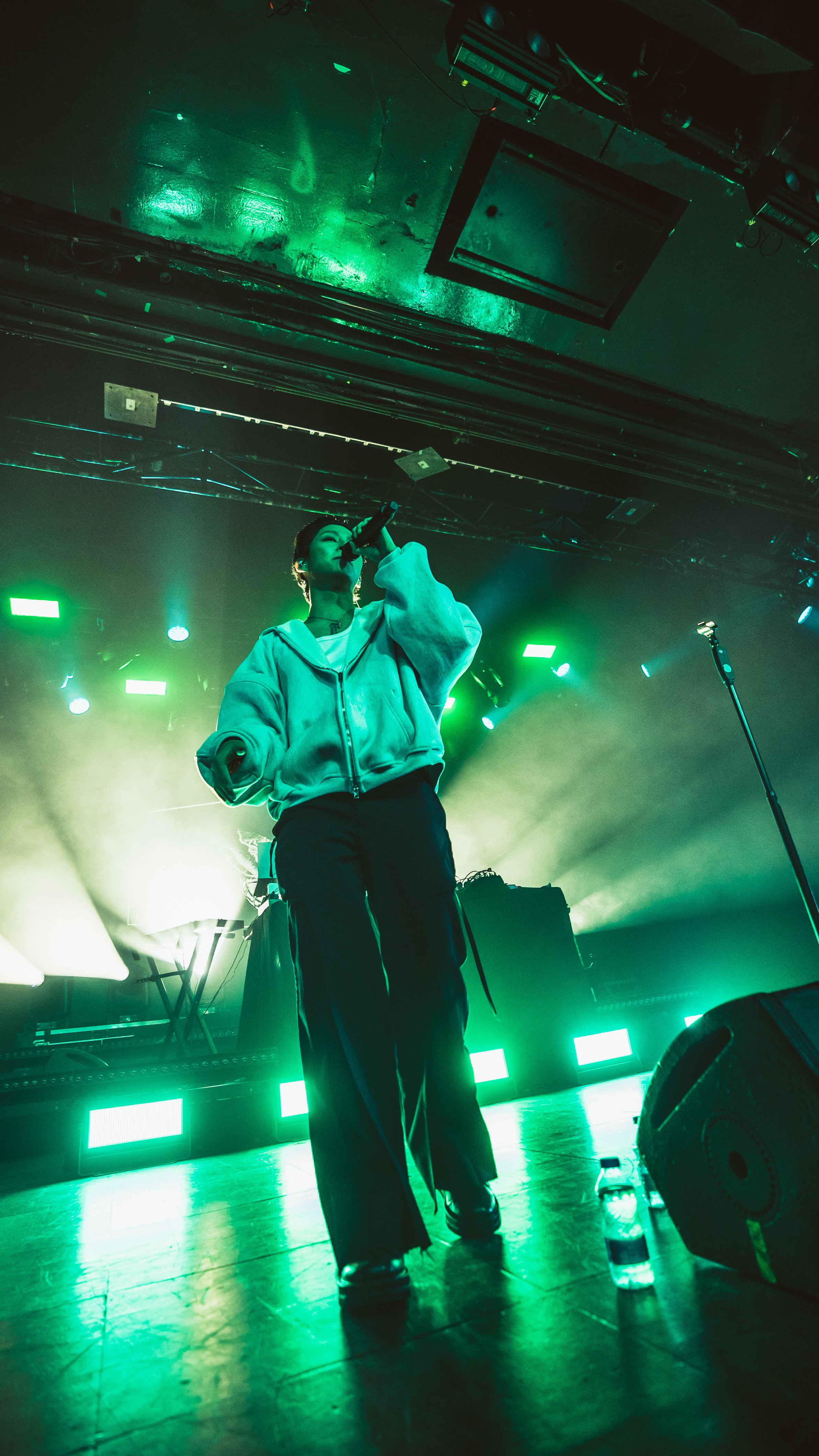 A female performer standing on stage with green lighting, wearing a hoodie and black pants, holding a microphone and singing. Stage equipment and a water bottle are visible in the foreground.